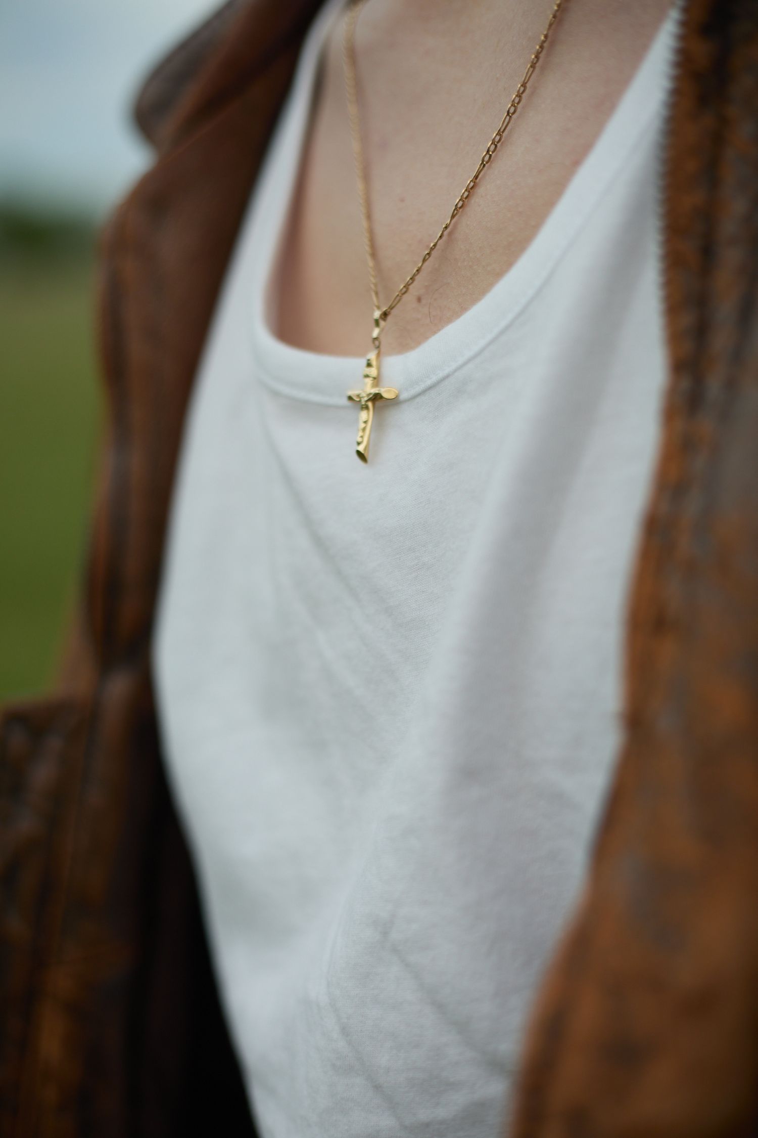Gold cross pendant necklace on a white t-shirt under a brown leather jacket outdoors.