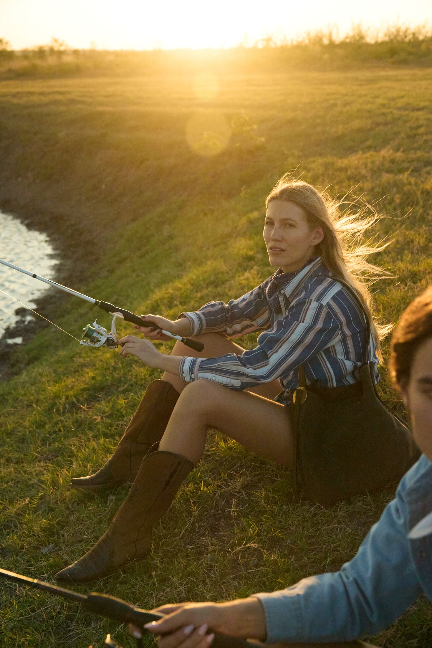 Blonde woman in striped shirt and brown boots holds a fishing rod by a river at golden sunset.