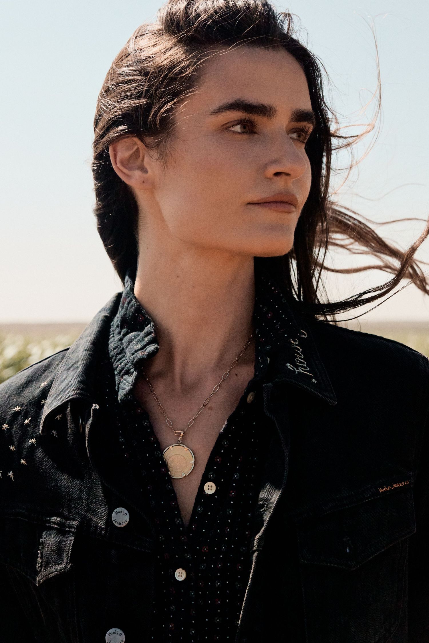 A woman with dark hair wearing a black denim jacket and gold pendant necklace gazes into the distance outdoors.