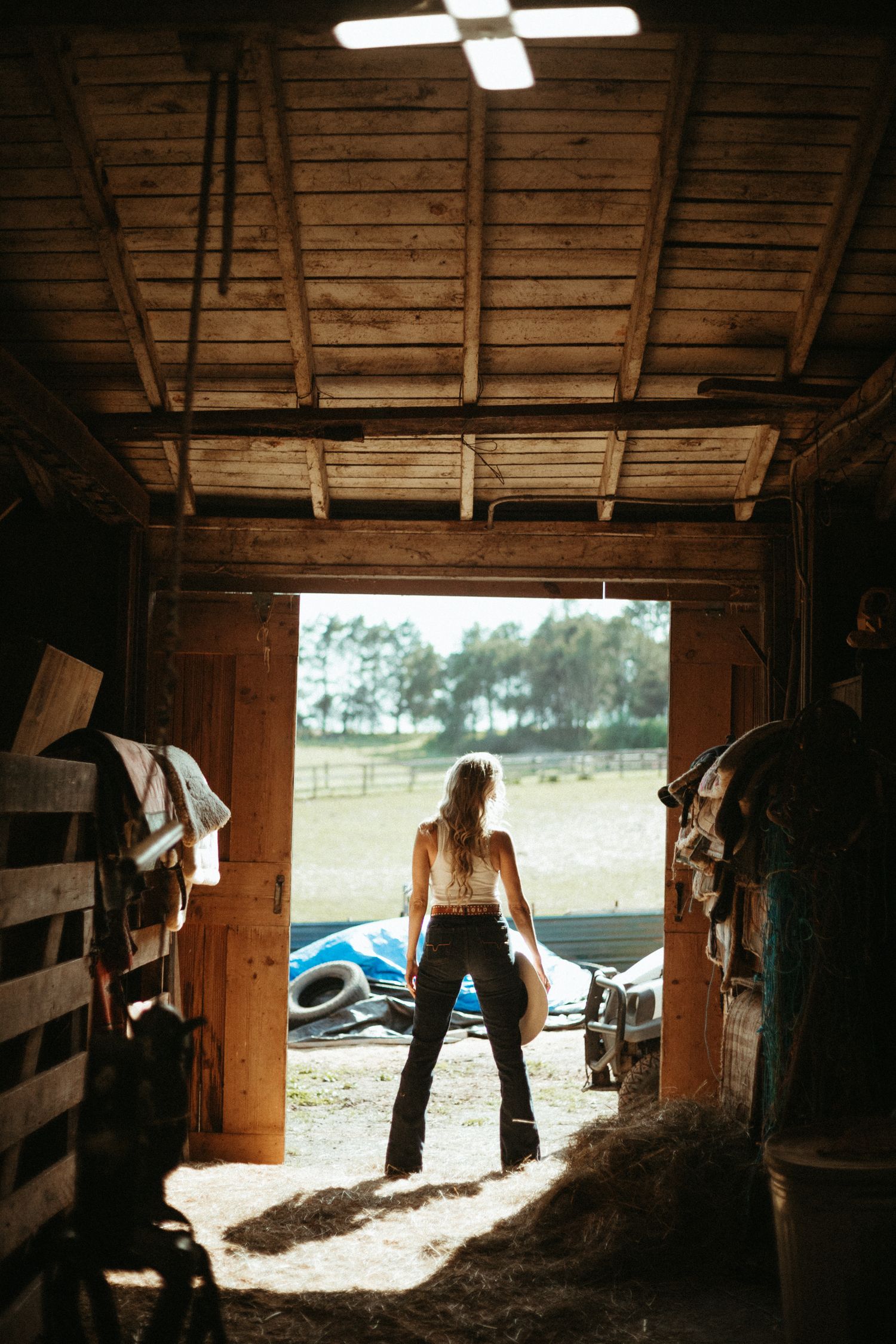 Woman in cowboy hat standing in barn doorway overlooking sunny green pasture.