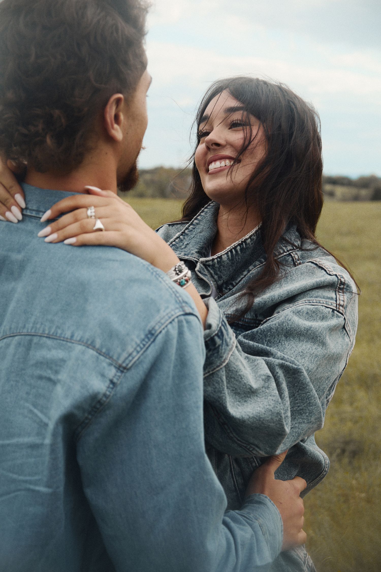 Happy couple embracing outdoors in denim jackets, smiling woman with dark hair in a green field.