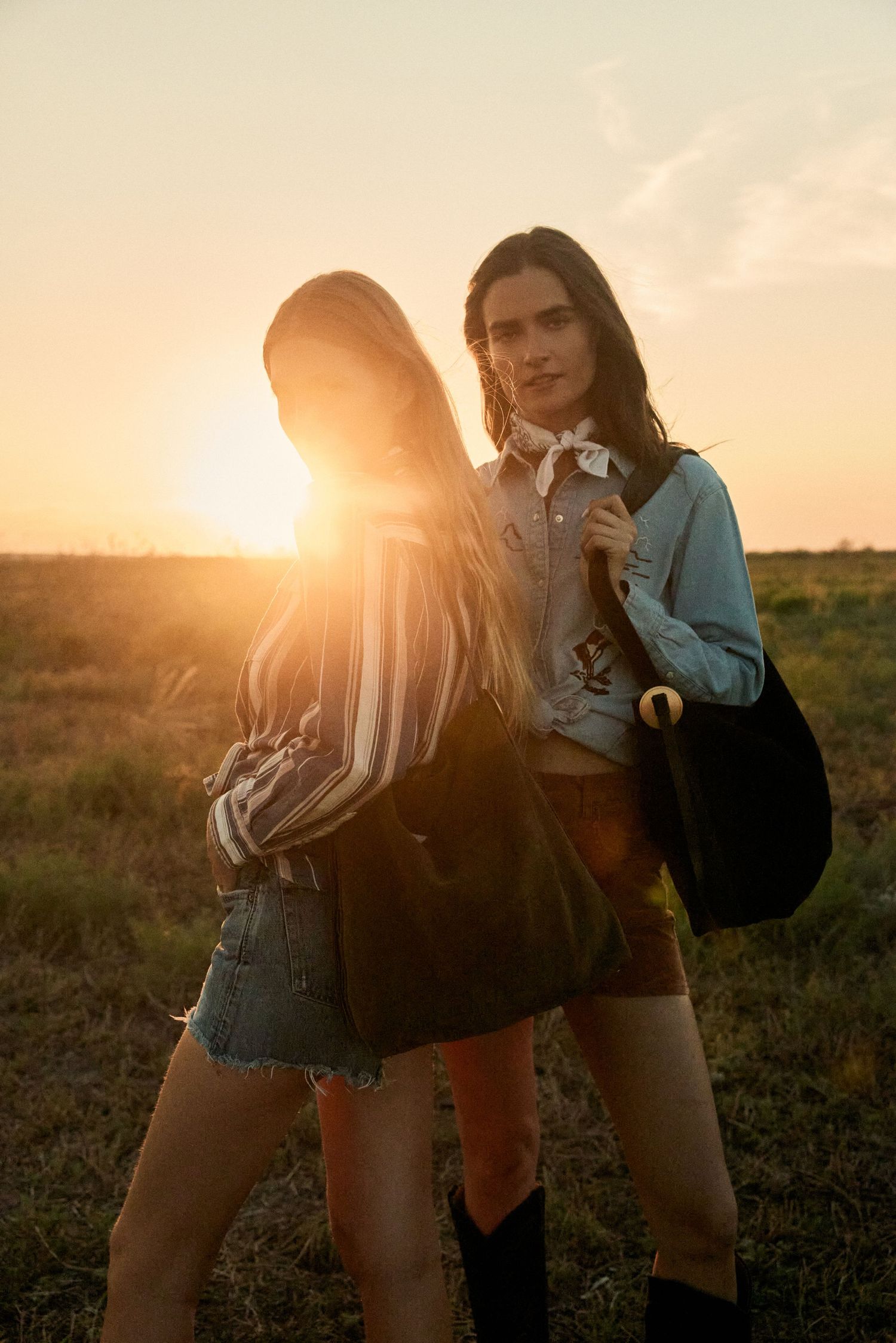 Two women in western boho outfits posing back to back in open field at sunset.