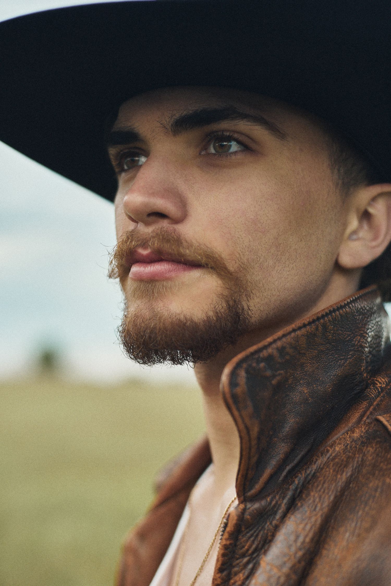Close-up portrait of a young man with beard wearing a black cowboy hat and brown leather jacket outdoors.