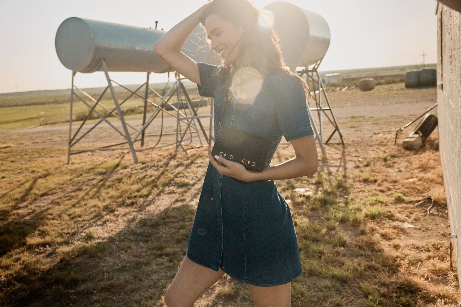 Young woman in denim mini skirt posing in sunlit rural field with farm equipment behind her.