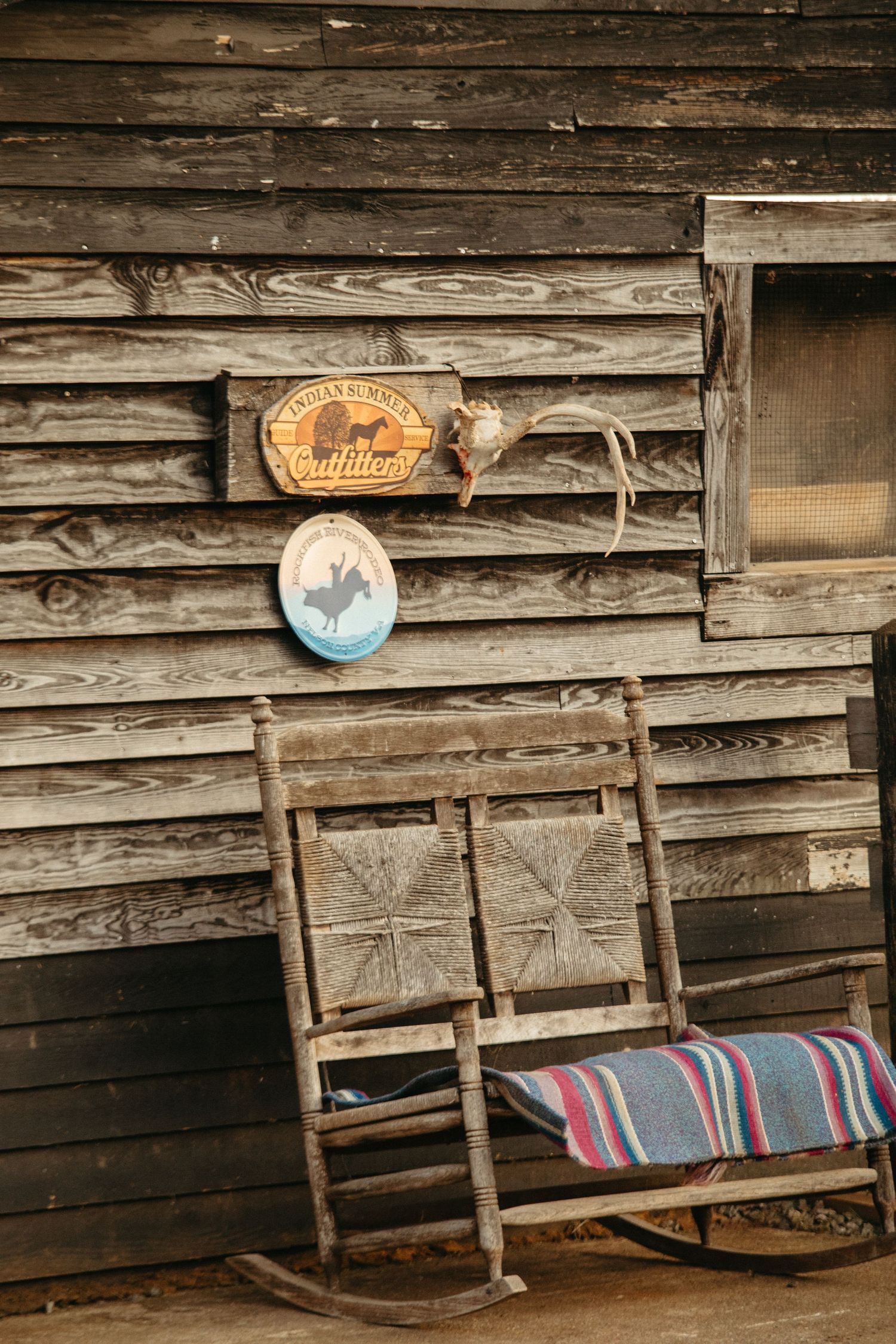 Vintage rocking chair with striped blanket beside rustic wooden barn wall with rodeo signs.