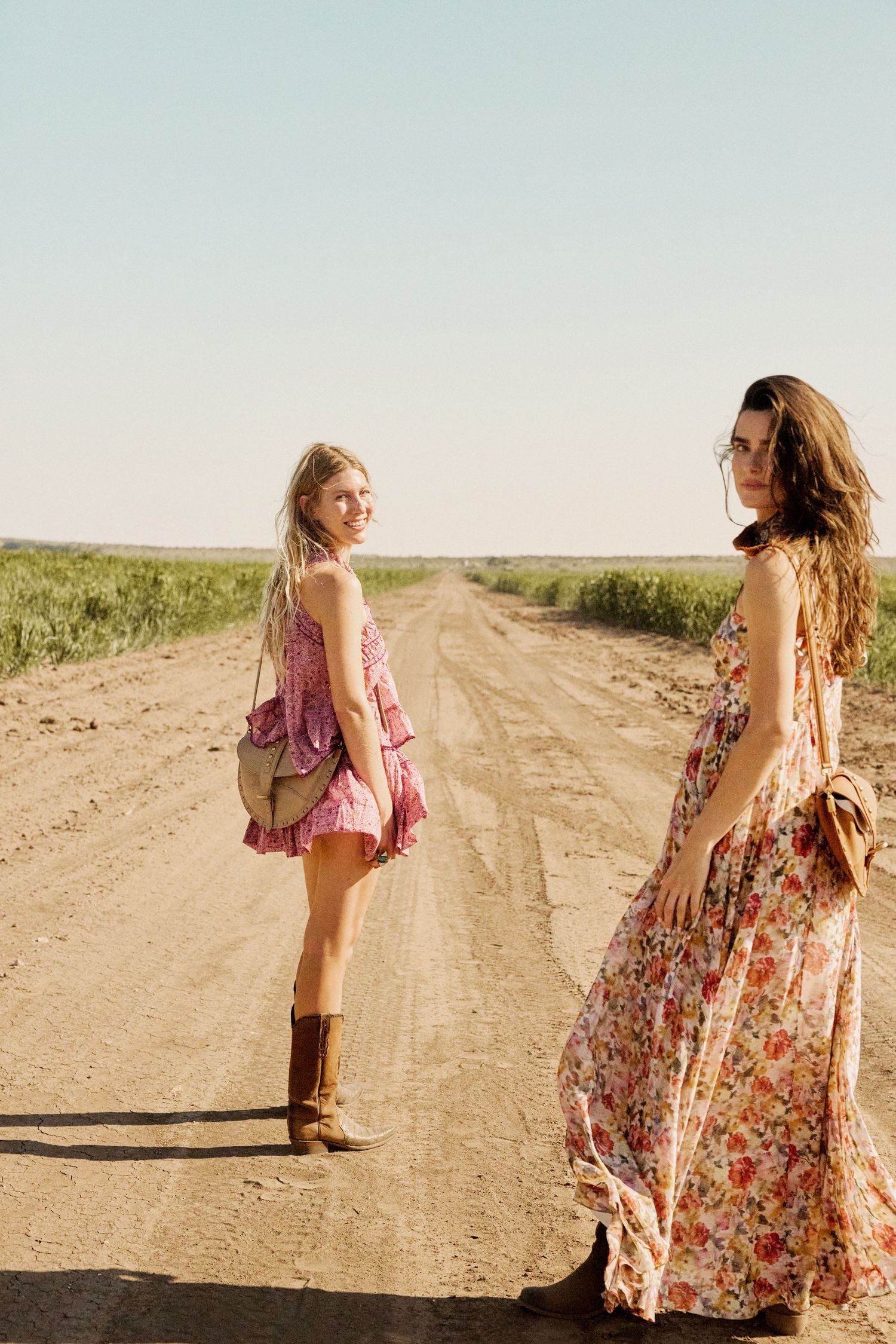 Two women in boho floral dresses posing on a sunny dirt road surrounded by fields.