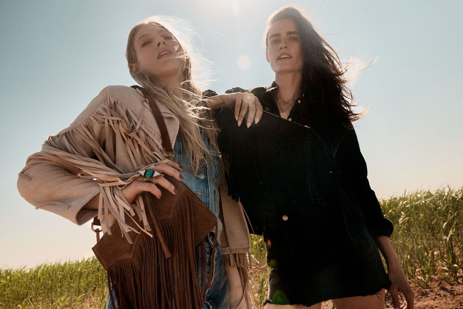 Two women in fringe and western-inspired outfits standing together in a bright sunny field.