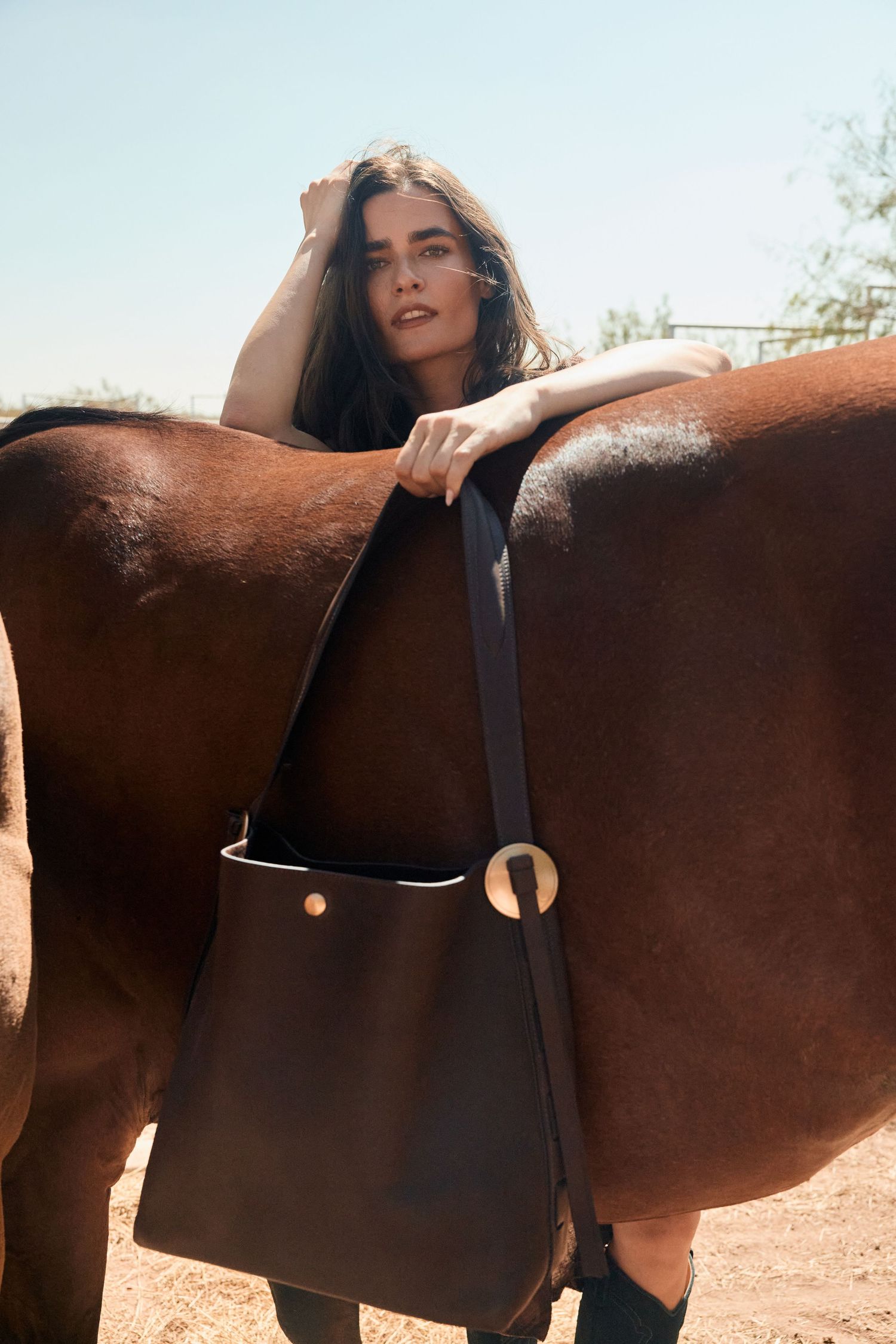Model in dark leather shoulder bag leans against brown horse outdoors in sunny setting.