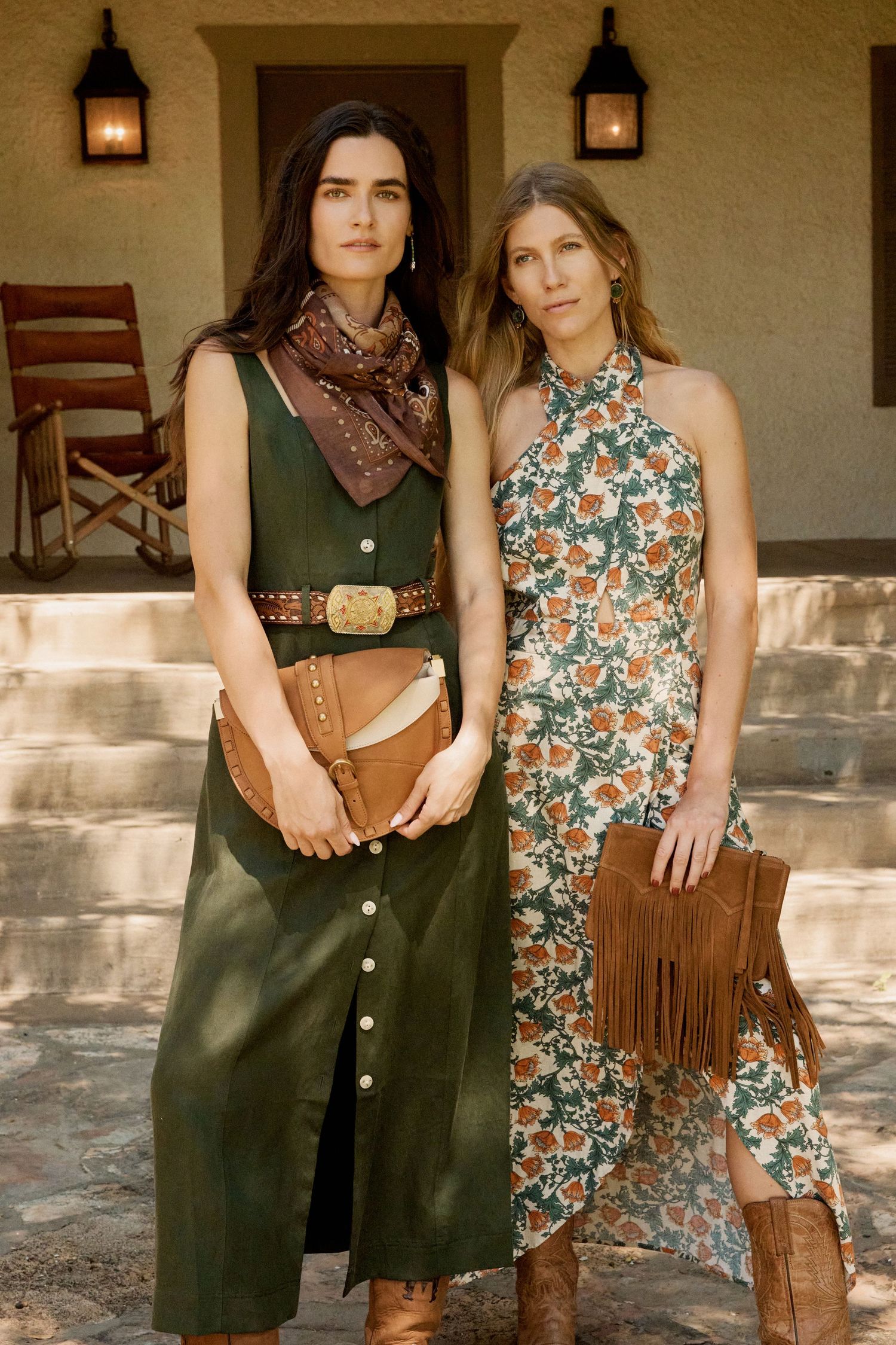 Two women in boho western outfits holding fringe bags outside a rustic adobe building.