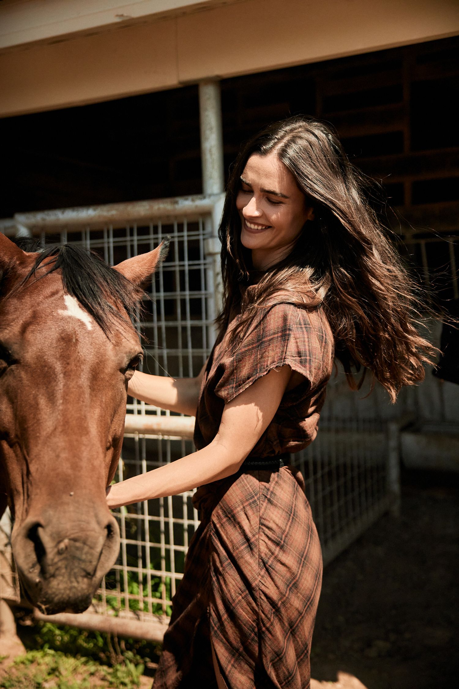 Smiling brunette woman in plaid brown dress gently pets a horse outside a stable in sunlight.