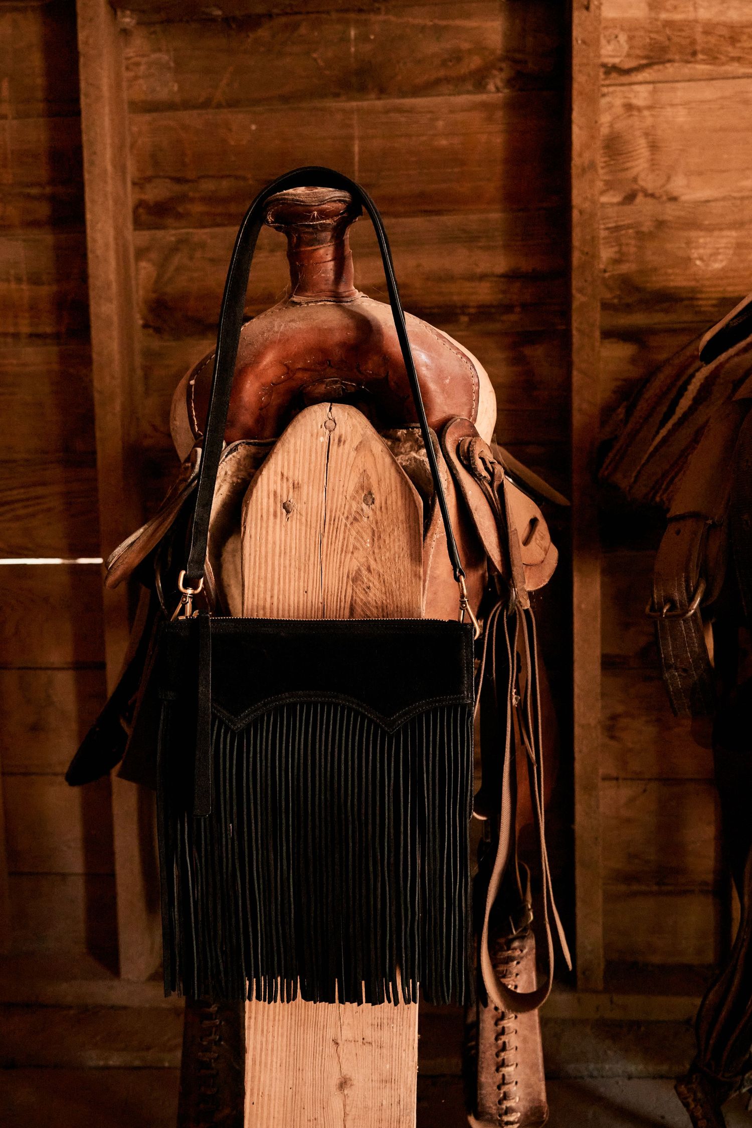 Vintage western leather saddle with black fringe bag displayed on wooden stand in barn.