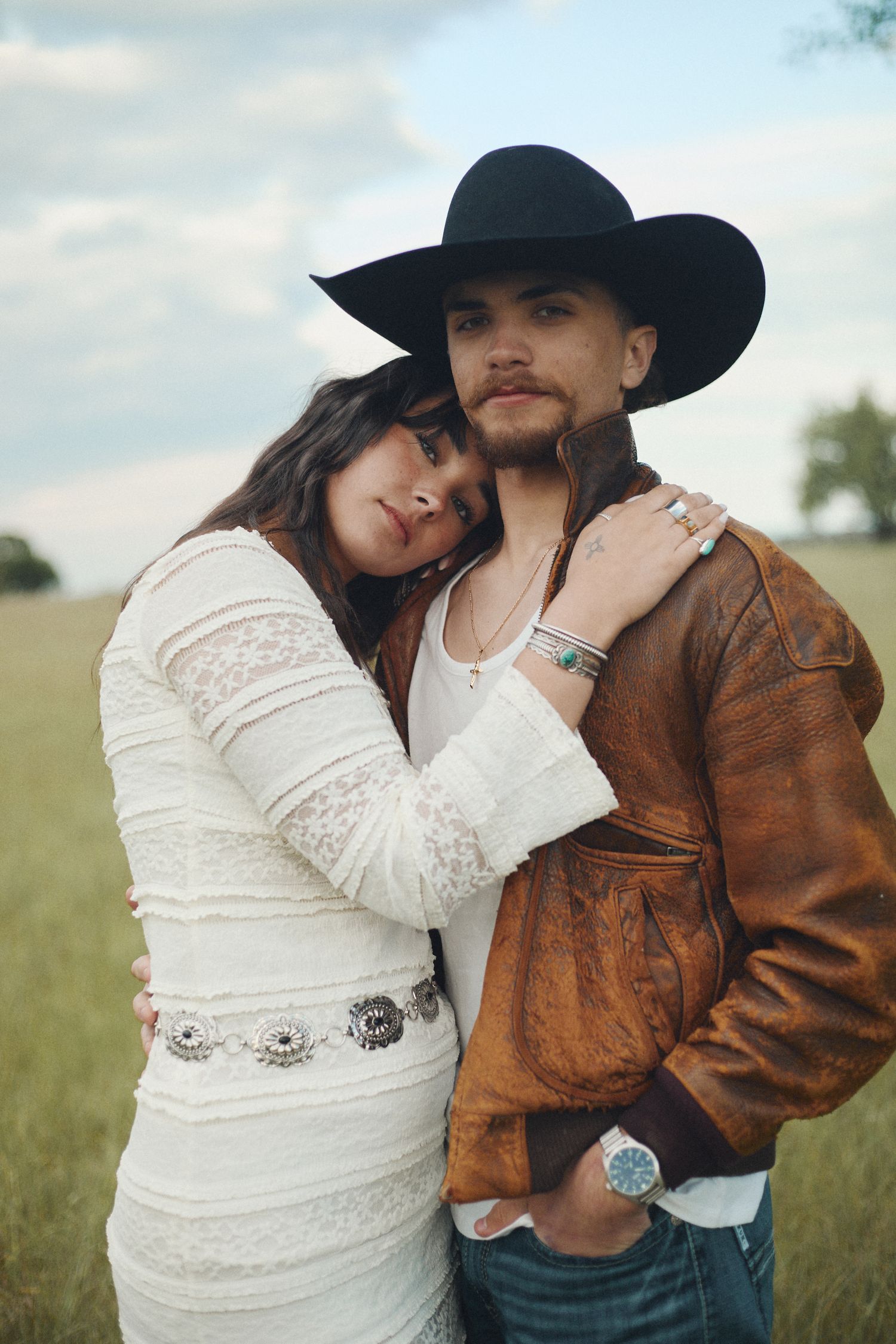 A couple embracing in a field, woman in white lace dress and man in black cowboy hat and brown leather jacket.