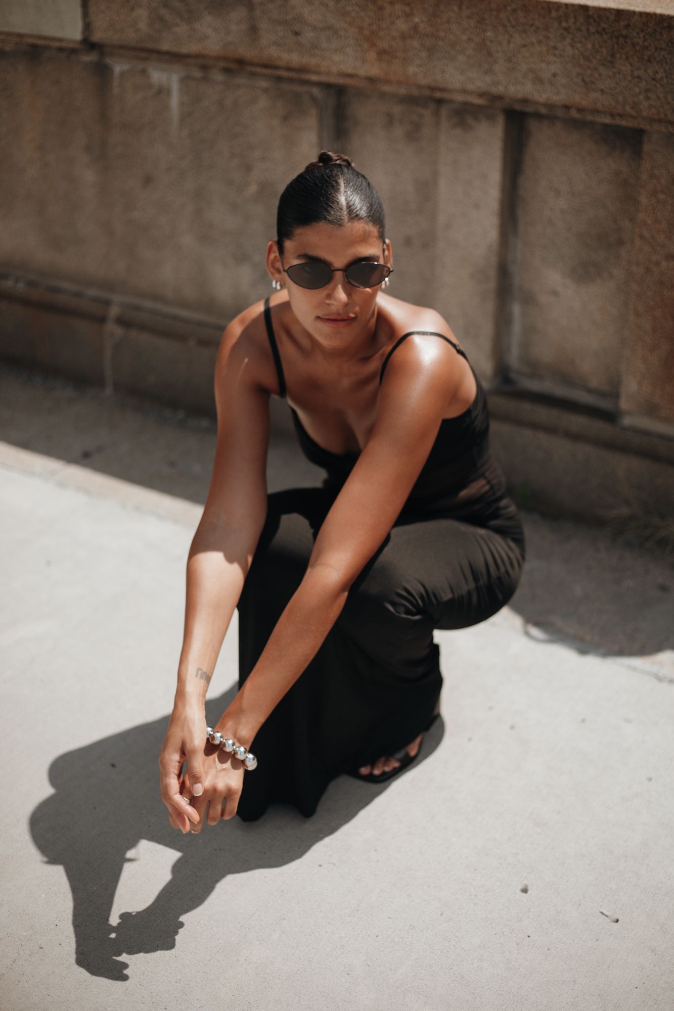 Woman in black outfit crouches on pavement wearing sunglasses and a pearl bracelet in bright sunlight.