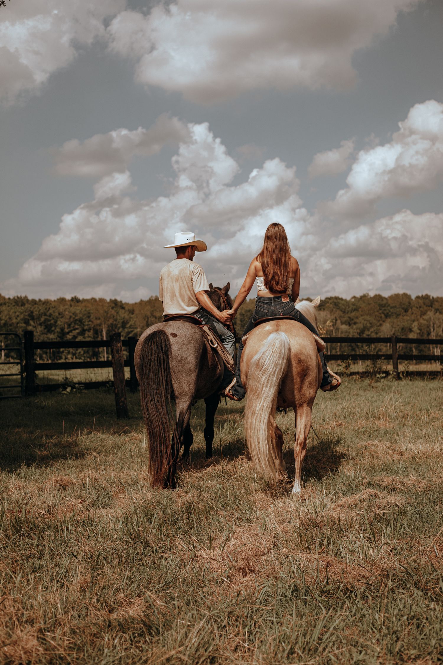 Couple riding horses side by side in a green pasture under a dramatic cloudy sky.