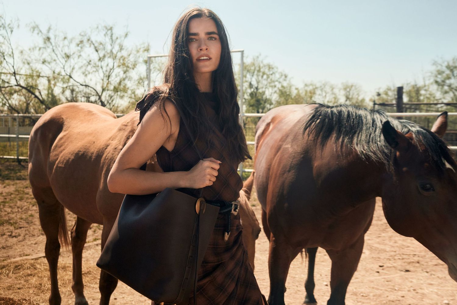 Woman in black dress standing between brown horses in a sunny outdoor ranch setting.