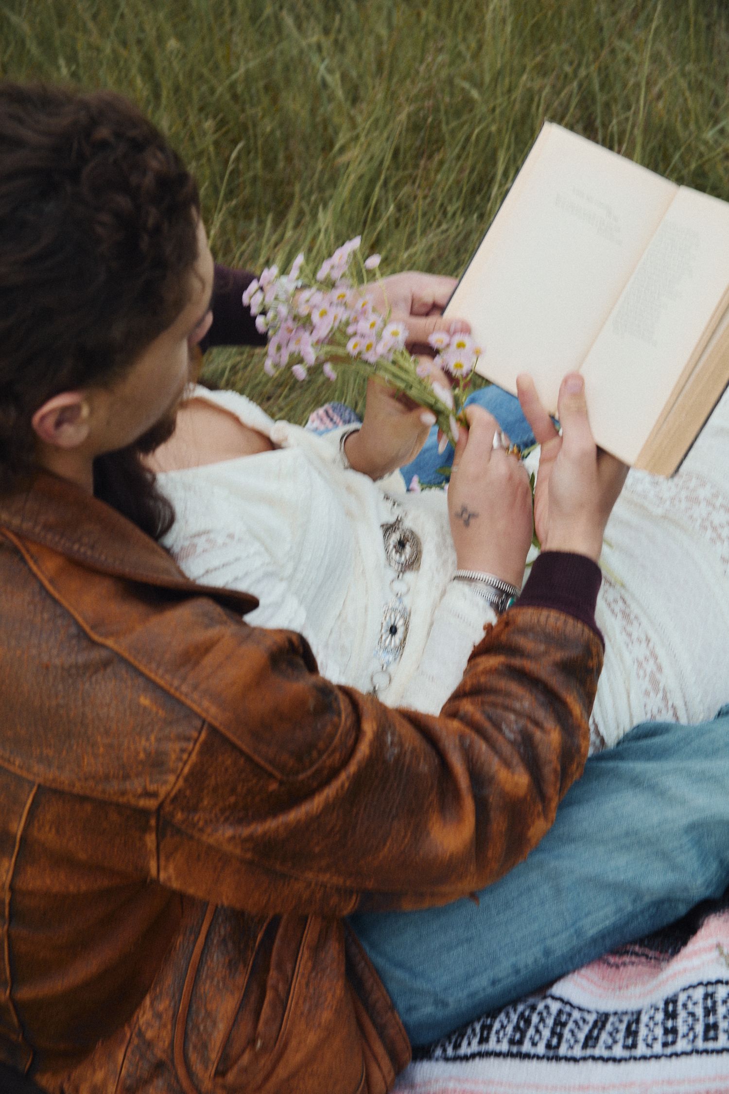 Couple relaxing outdoors in a field, reading a book together while holding wildflowers, wearing bohemian style clothing.
