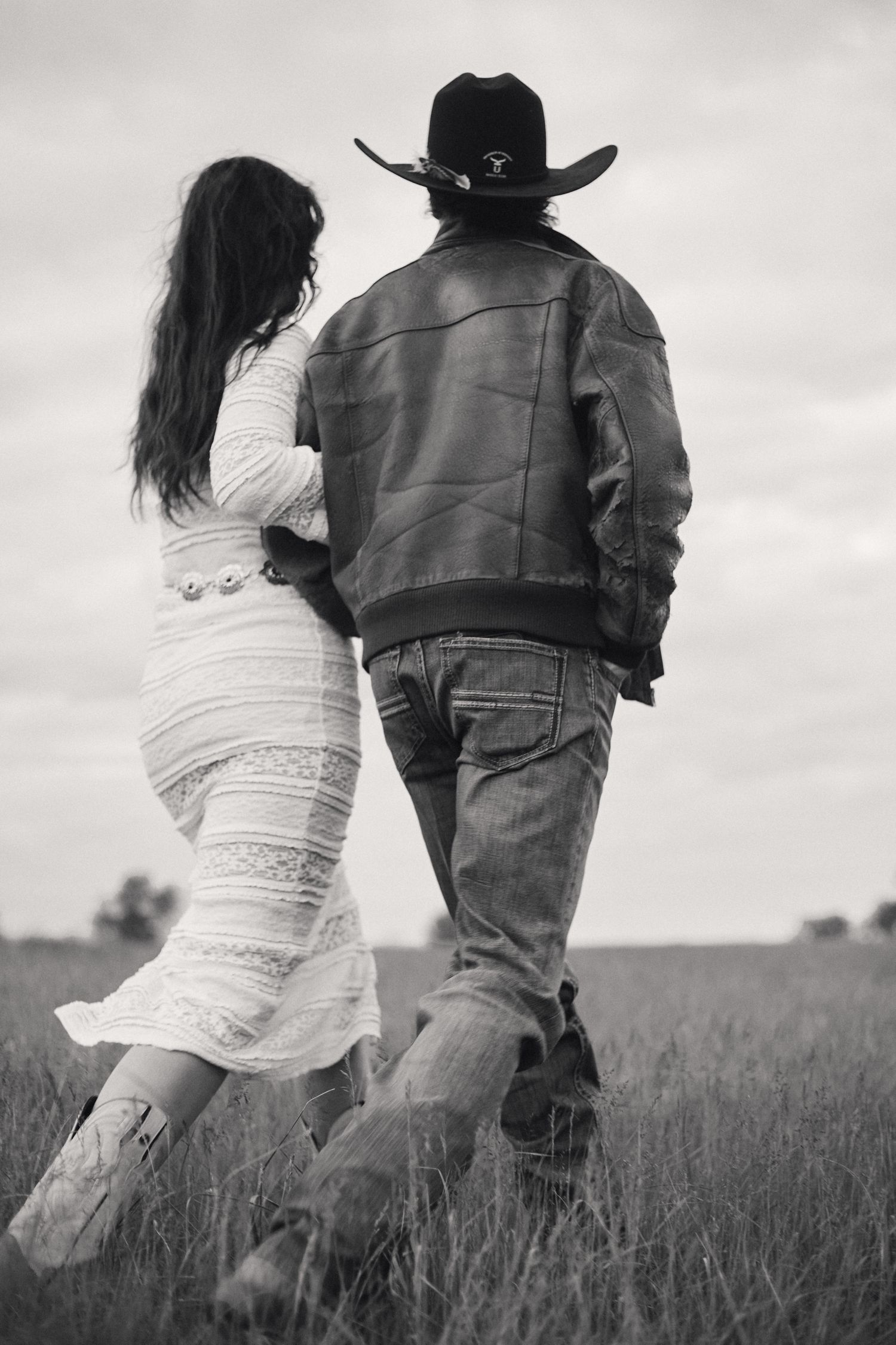 Black and white photo of a pregnant couple walking through a field, man wearing cowboy hat and leather jacket.
