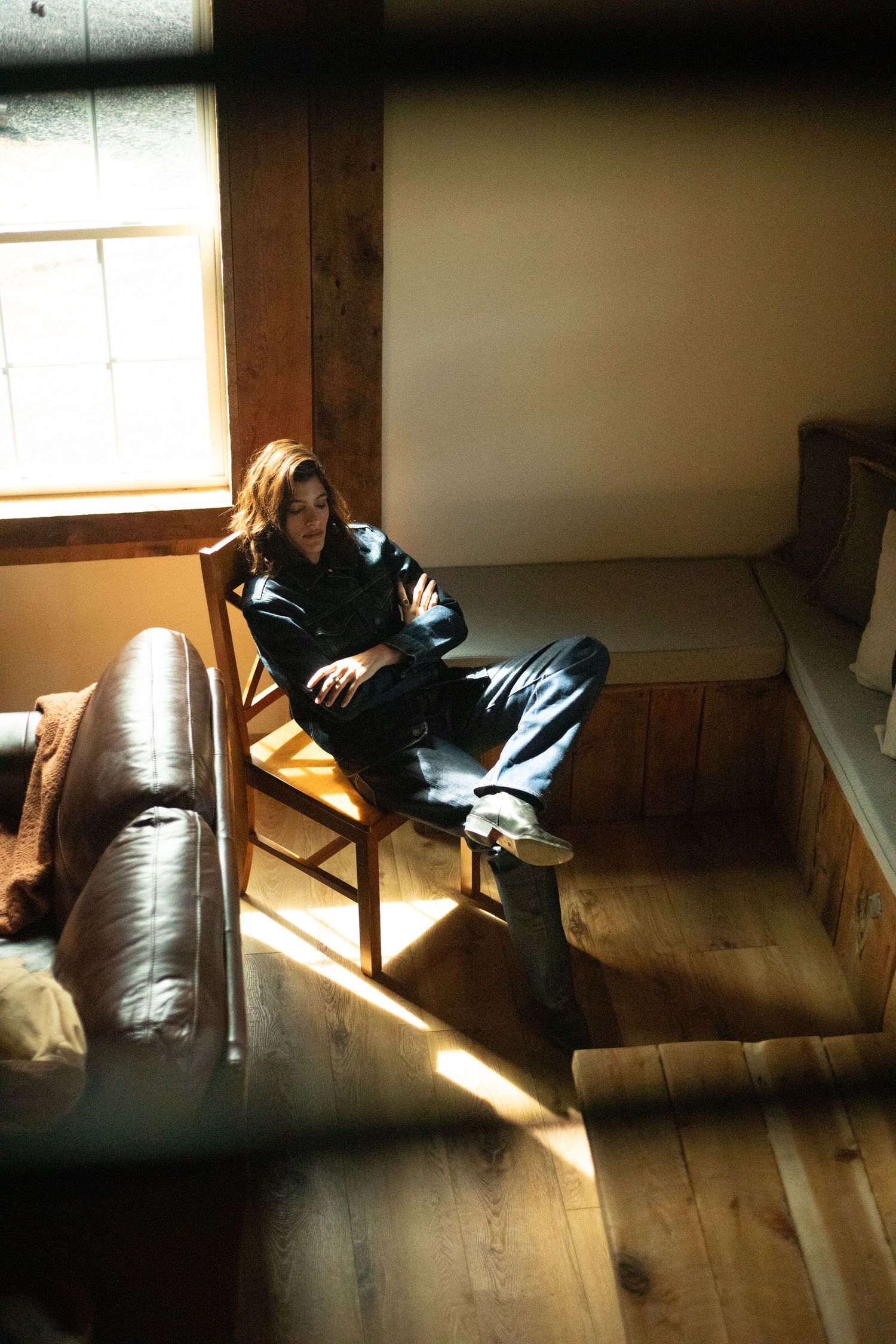 Person in navy outfit relaxes in wooden chair by sunlit window indoors.