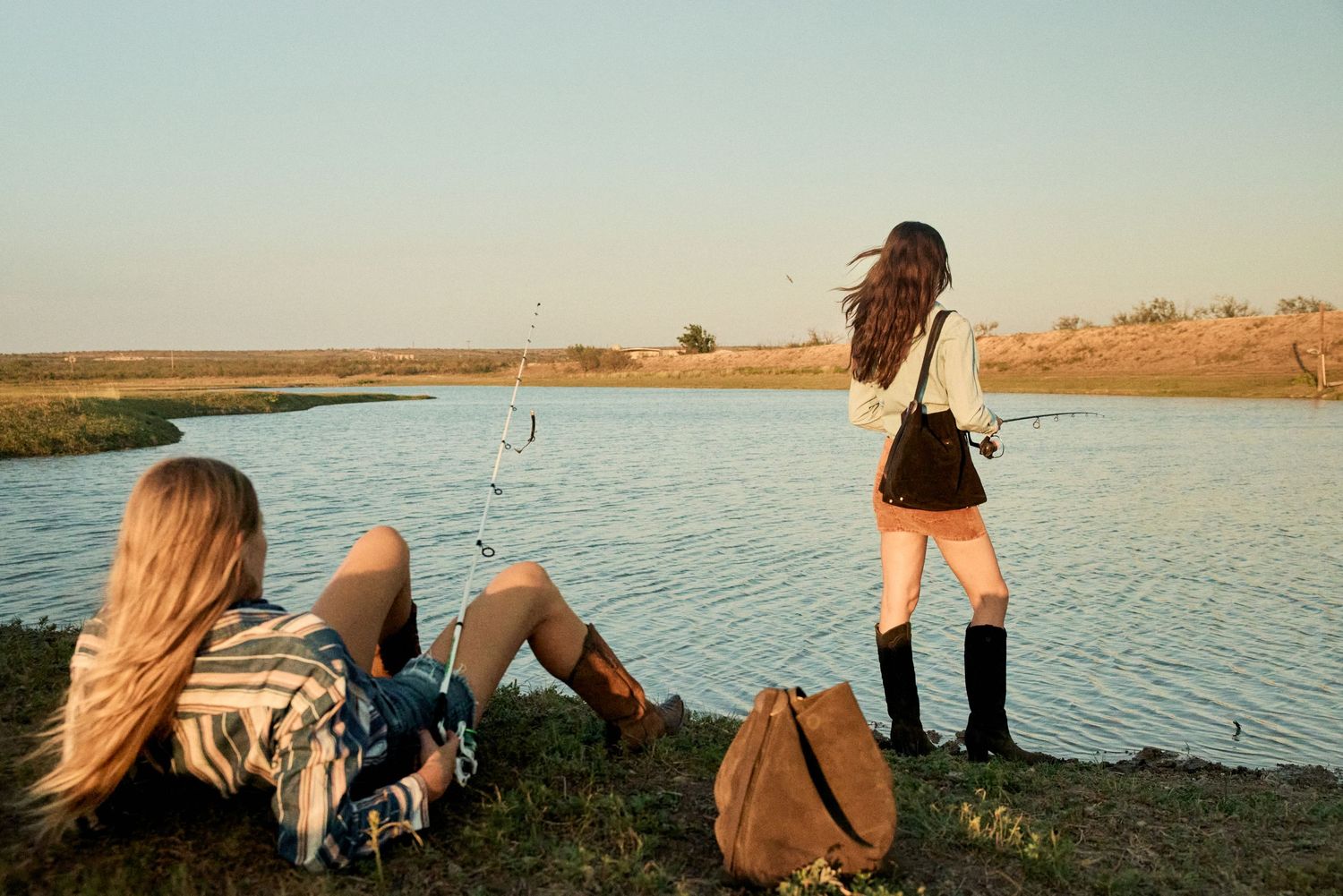 Two women fishing by a scenic lake at sunset, one sitting on grass, one standing with a fishing rod.