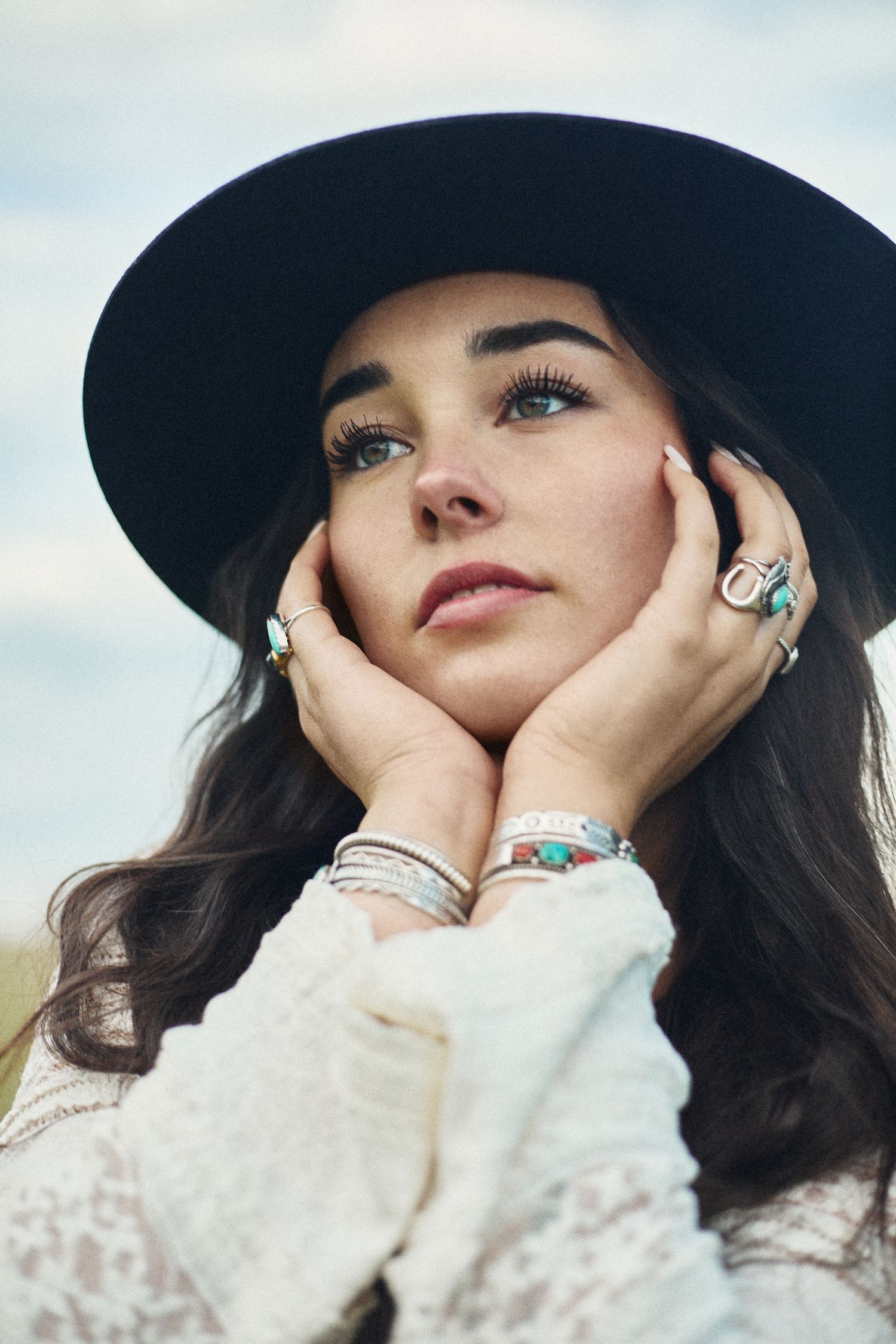 Young woman in black wide-brim hat with turquoise jewelry, hands framing her face, gazing upward outdoors.