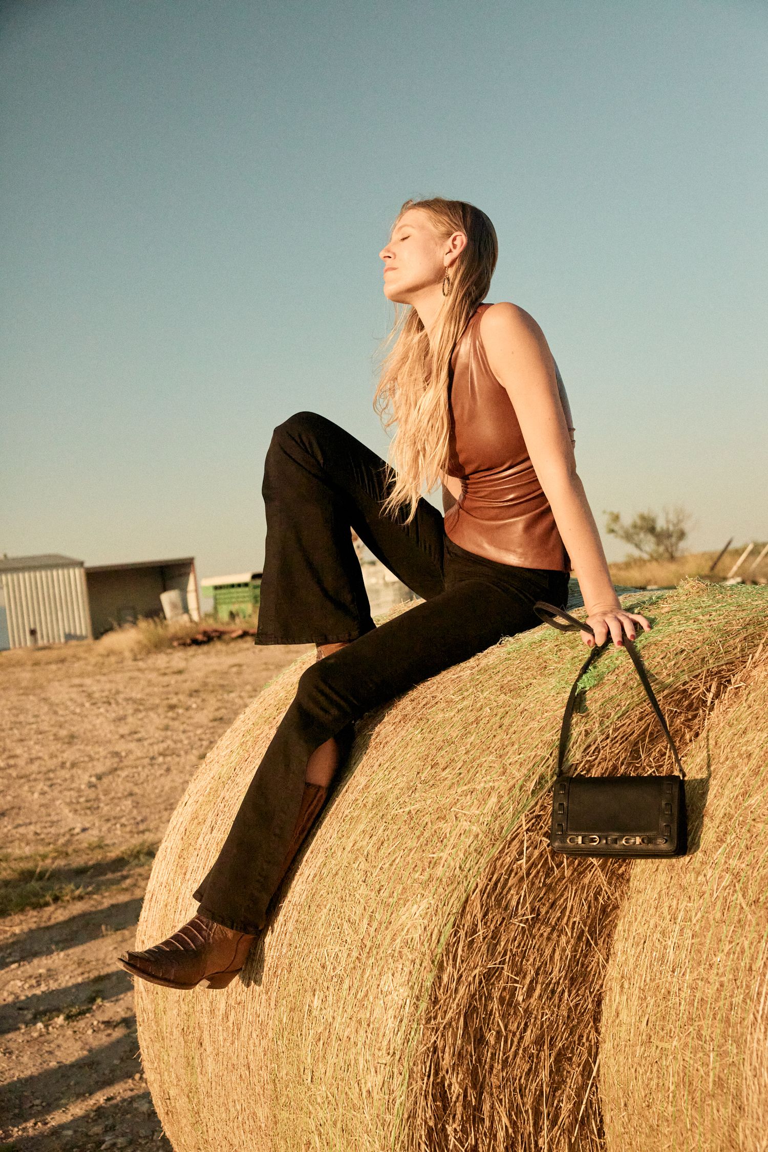 Blonde model in brown top and black pants sits atop a large hay bale holding a black handbag.