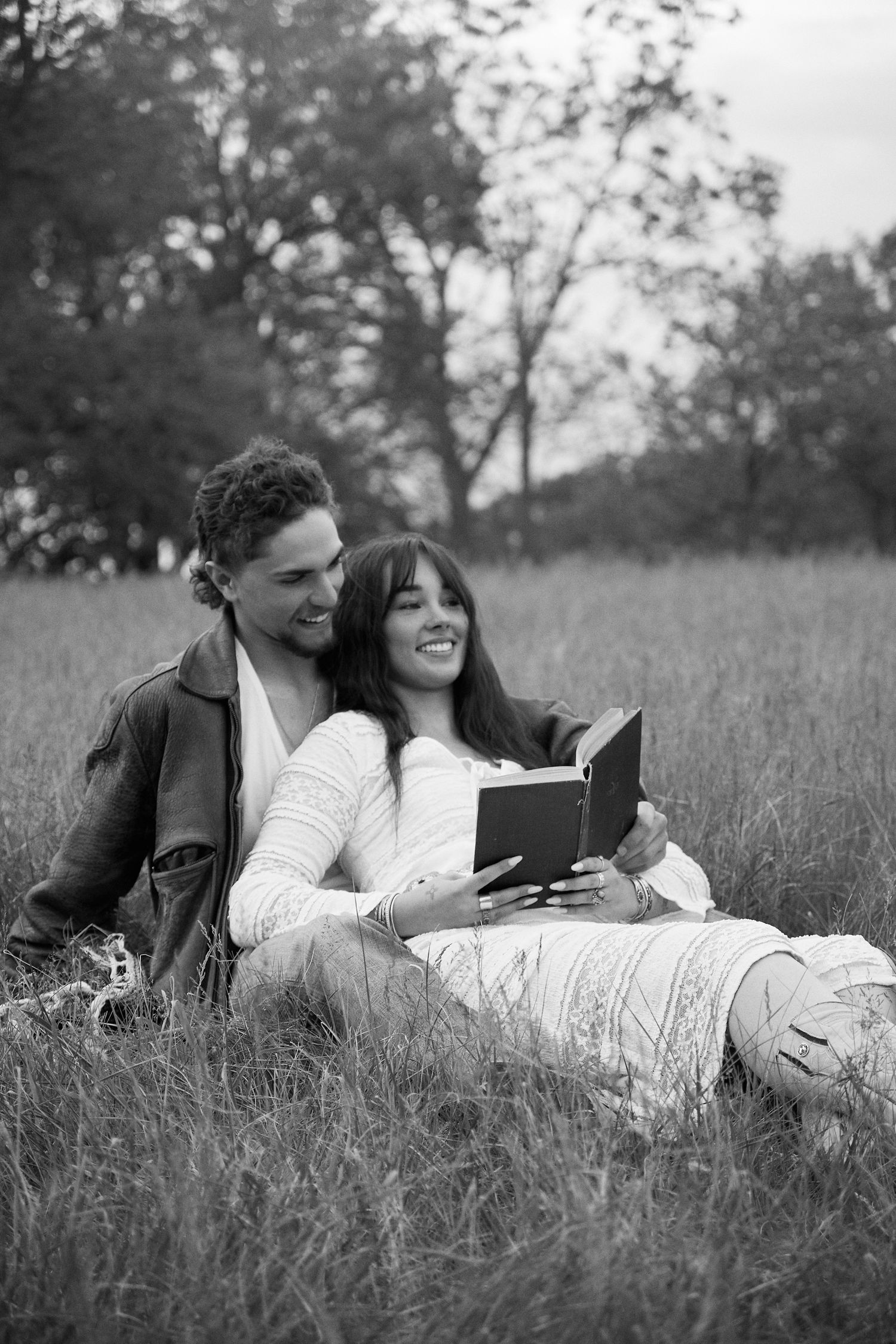 Black and white photo of a smiling couple sitting in a grassy field together, reading a book outdoors.