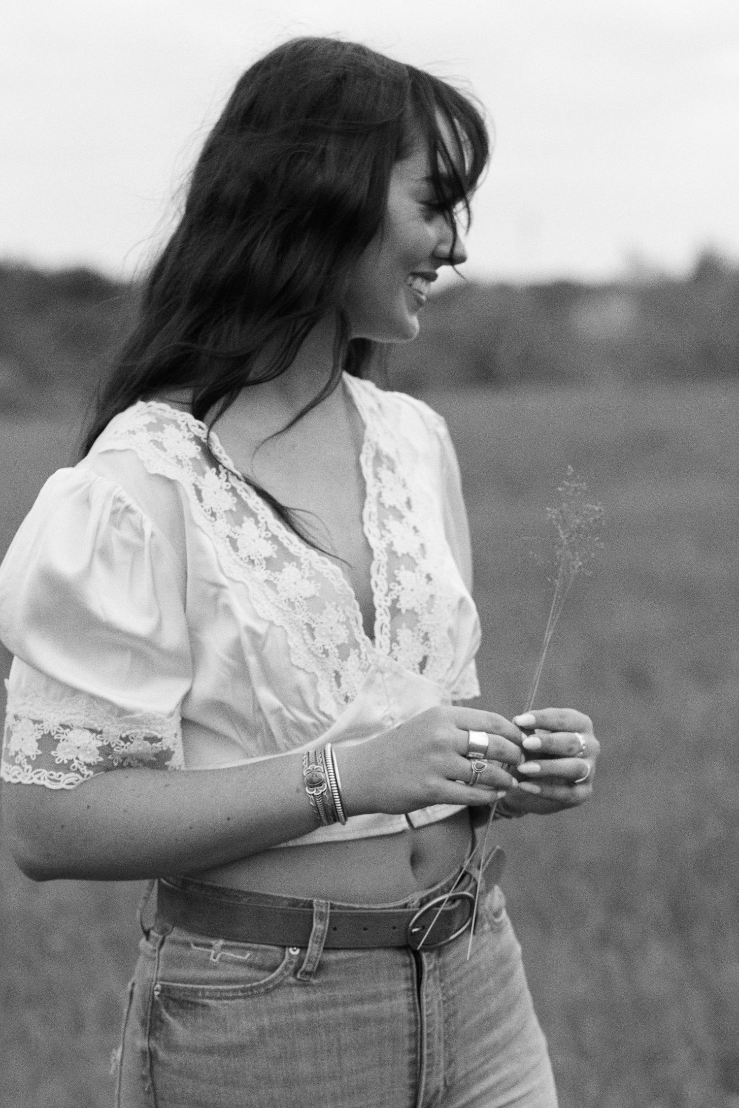 Black and white photo of a smiling woman in a white lace crop top and jeans holding a wildflower in a field.