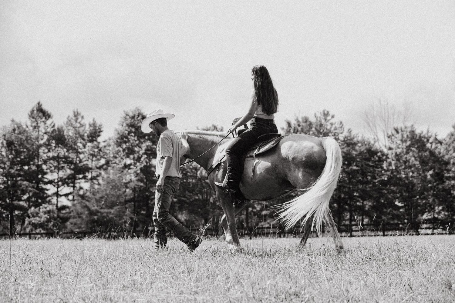 Black and white photo of a cowboy leading a woman on horseback through an open field with trees in background.