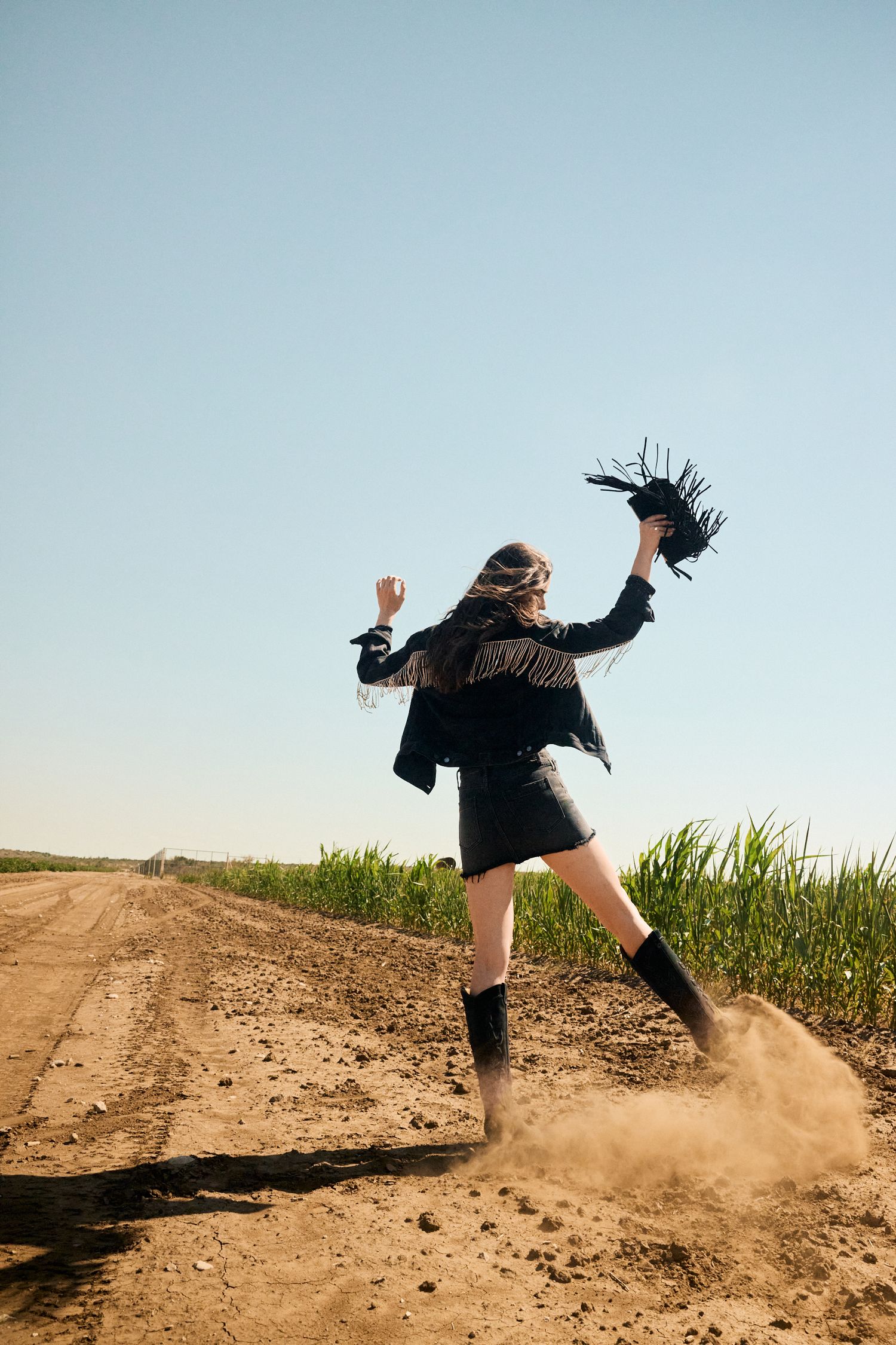 Woman in black fringe jacket and mini skirt jumps on a dusty dirt road beside a green field.