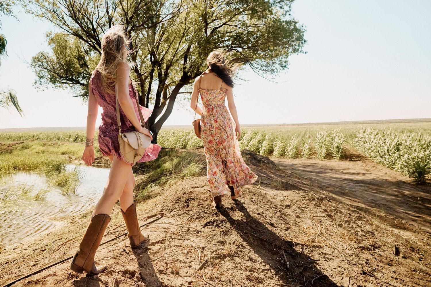 Two women in boho floral dresses walking along a rural dirt path near a field.