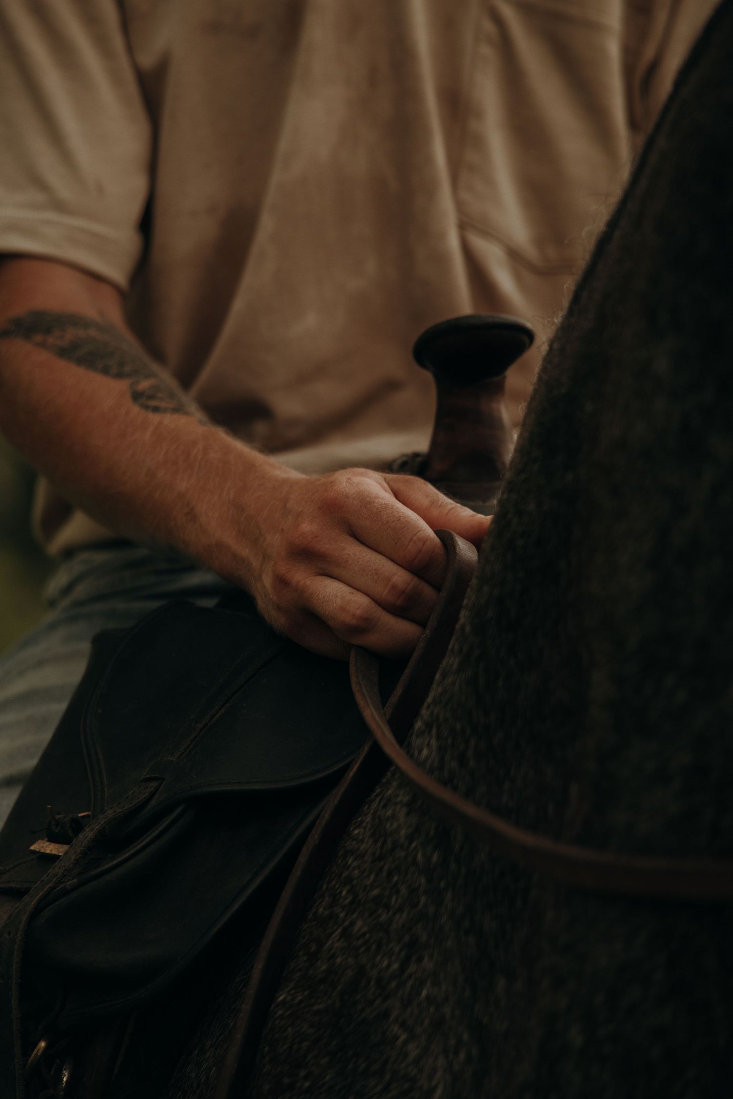Close-up of a tattooed cowboy hand gripping a leather saddle horn on a dark horse.