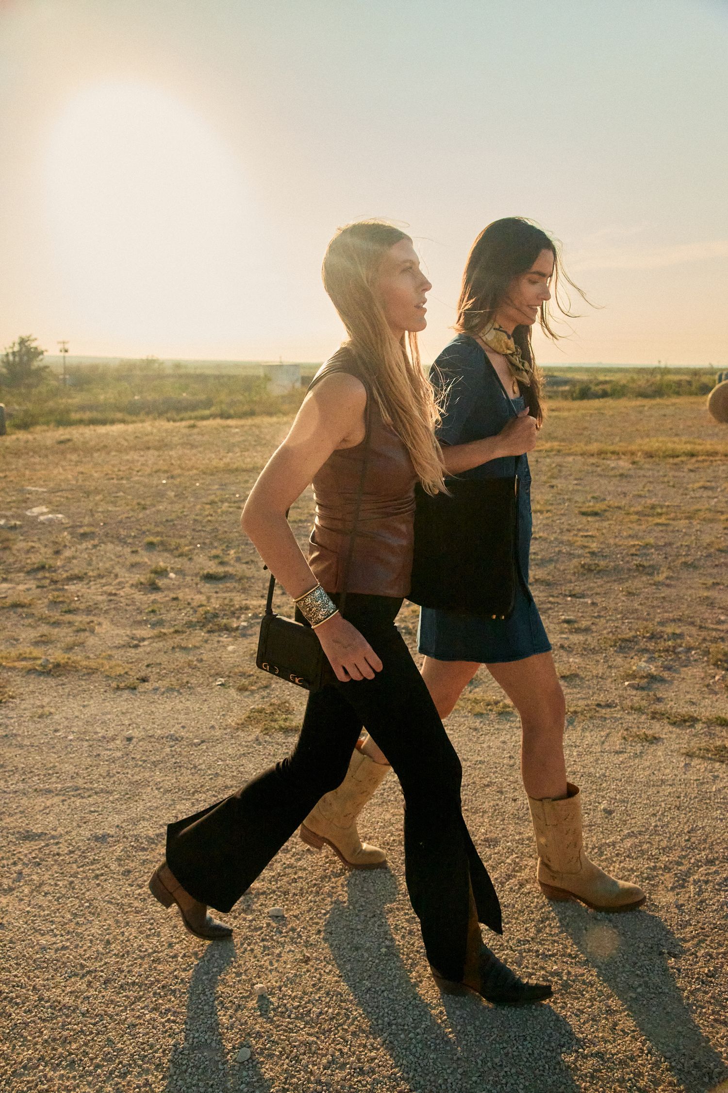 Two women in western-style outfits walk through a sunlit desert landscape wearing boots and carrying handbags.