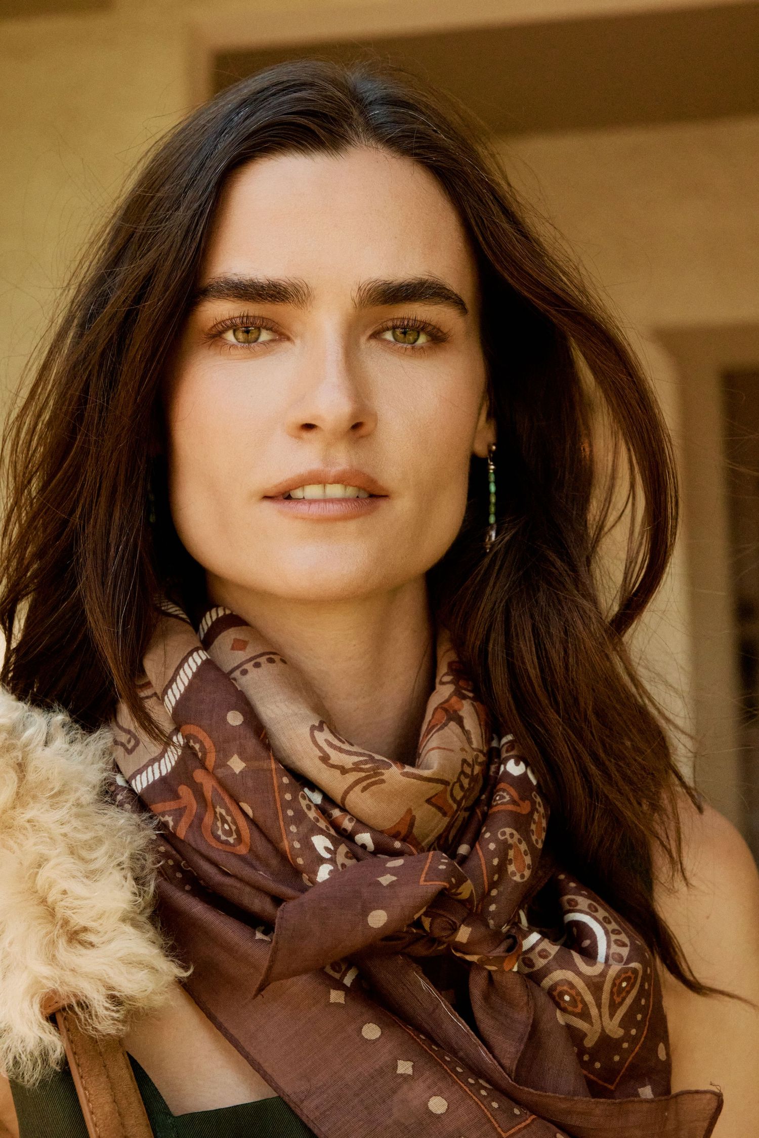 A brunette woman with green eyes wearing a brown paisley scarf and gold earrings outdoors.