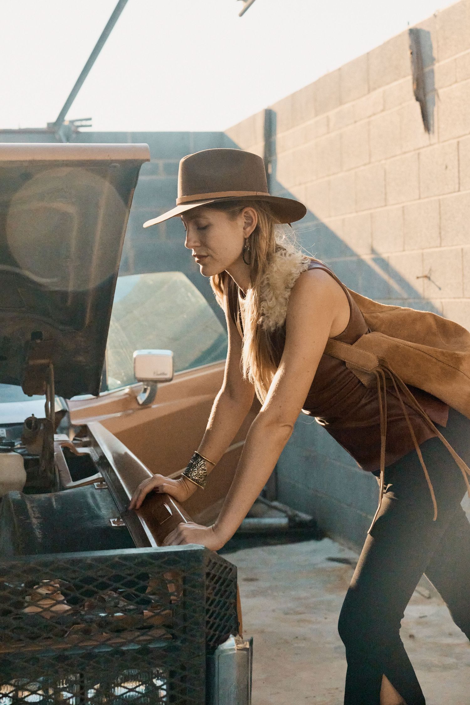 Woman in brown hat and fringe vest leaning over vintage truck bed in rustic outdoor setting.