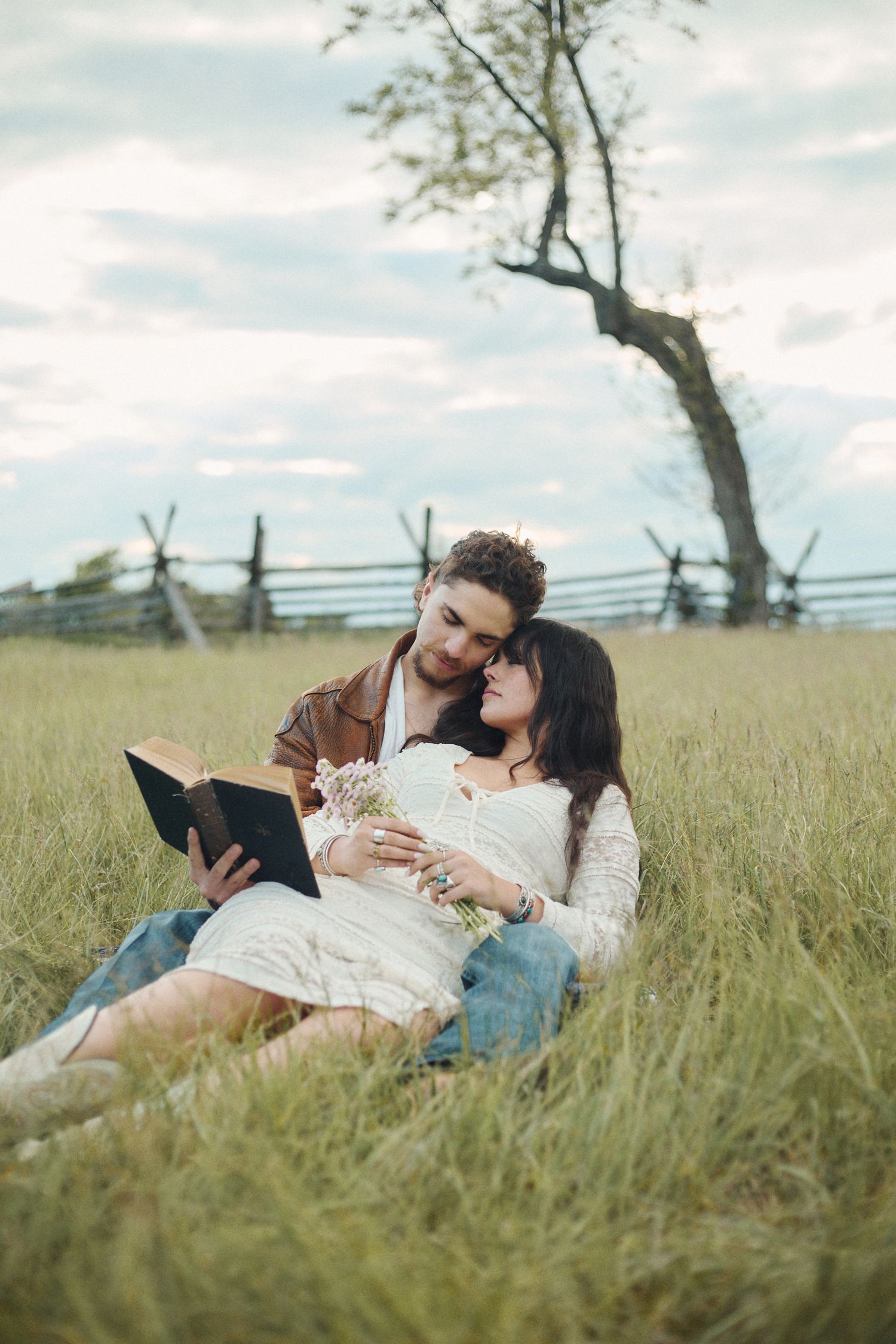 Romantic couple sitting in a grassy field, sharing a book and tender moment near a rustic fence.