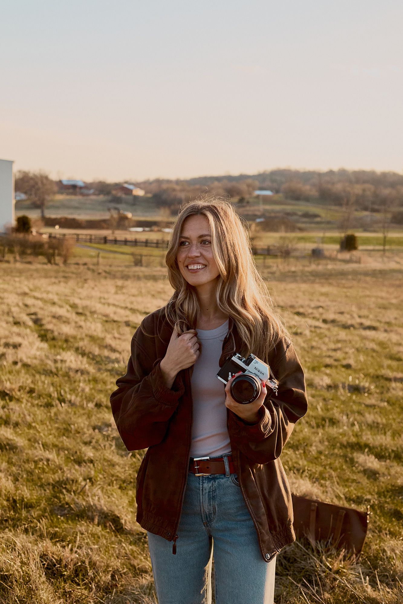 Smiling blonde woman holding a film camera in a sunny rural field wearing a brown jacket and jeans.