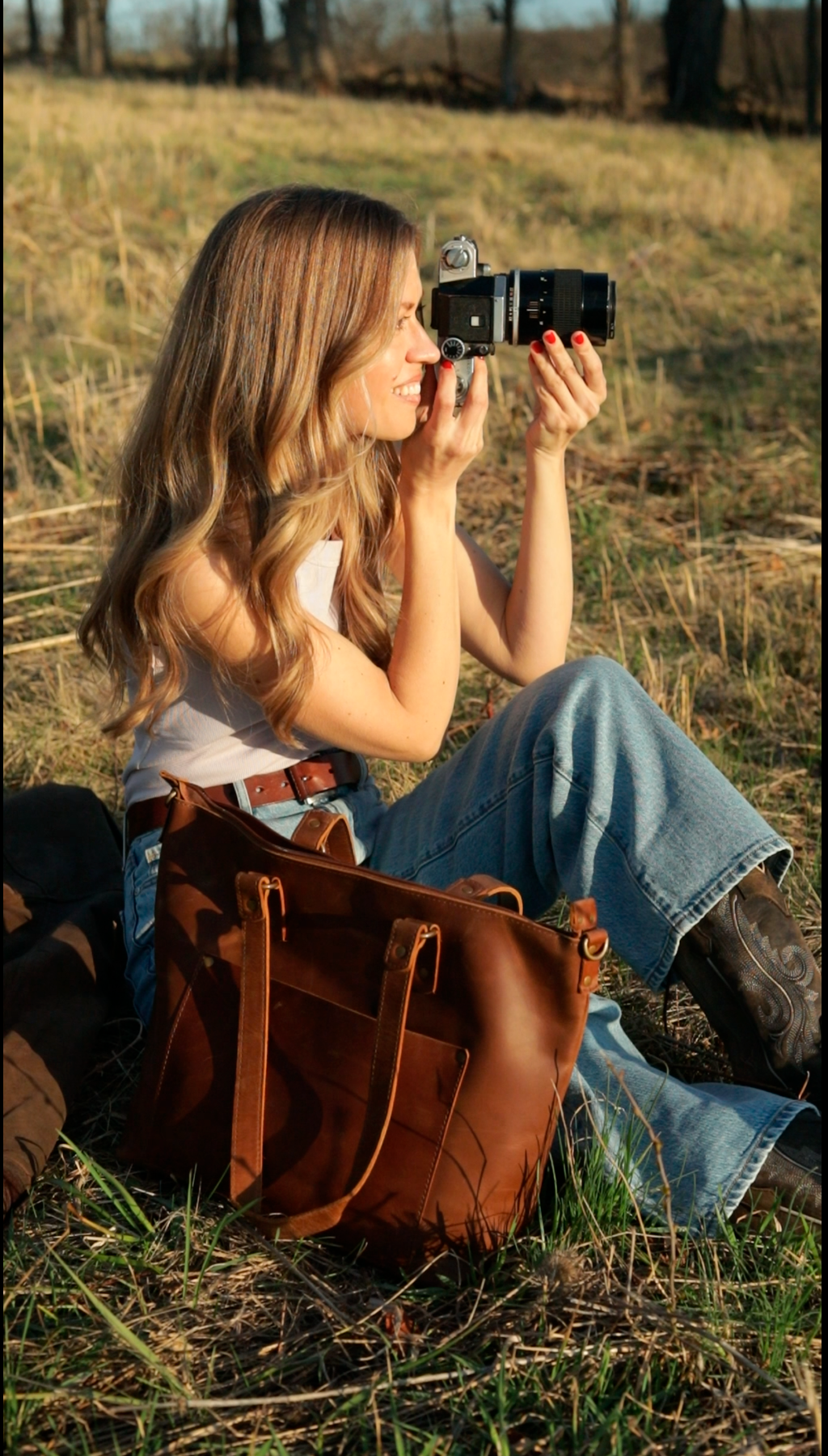Blonde woman sitting in a sunny field, holding a vintage camera beside a brown leather tote bag.