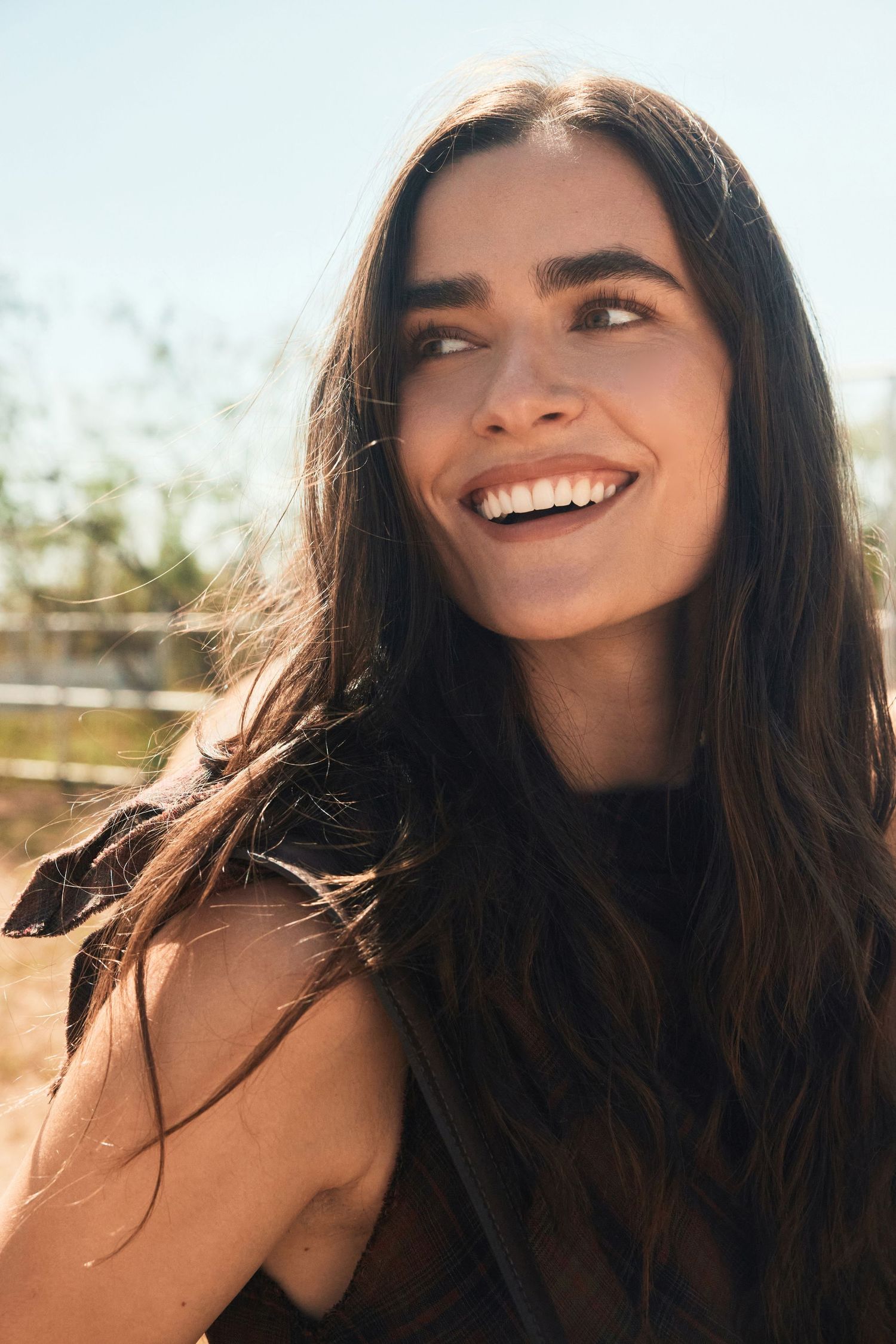 Smiling young woman with long dark hair outdoors in bright sunlight at a ranch.