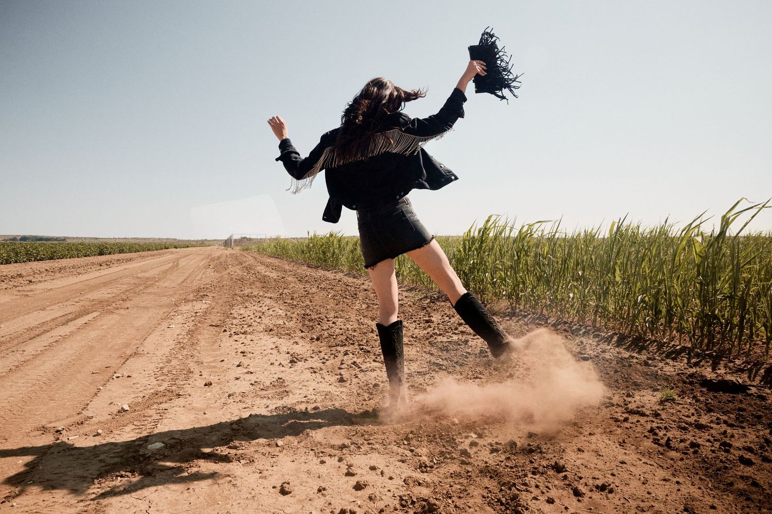 Woman in black outfit and boots jumping on a dusty dirt road beside green fields.