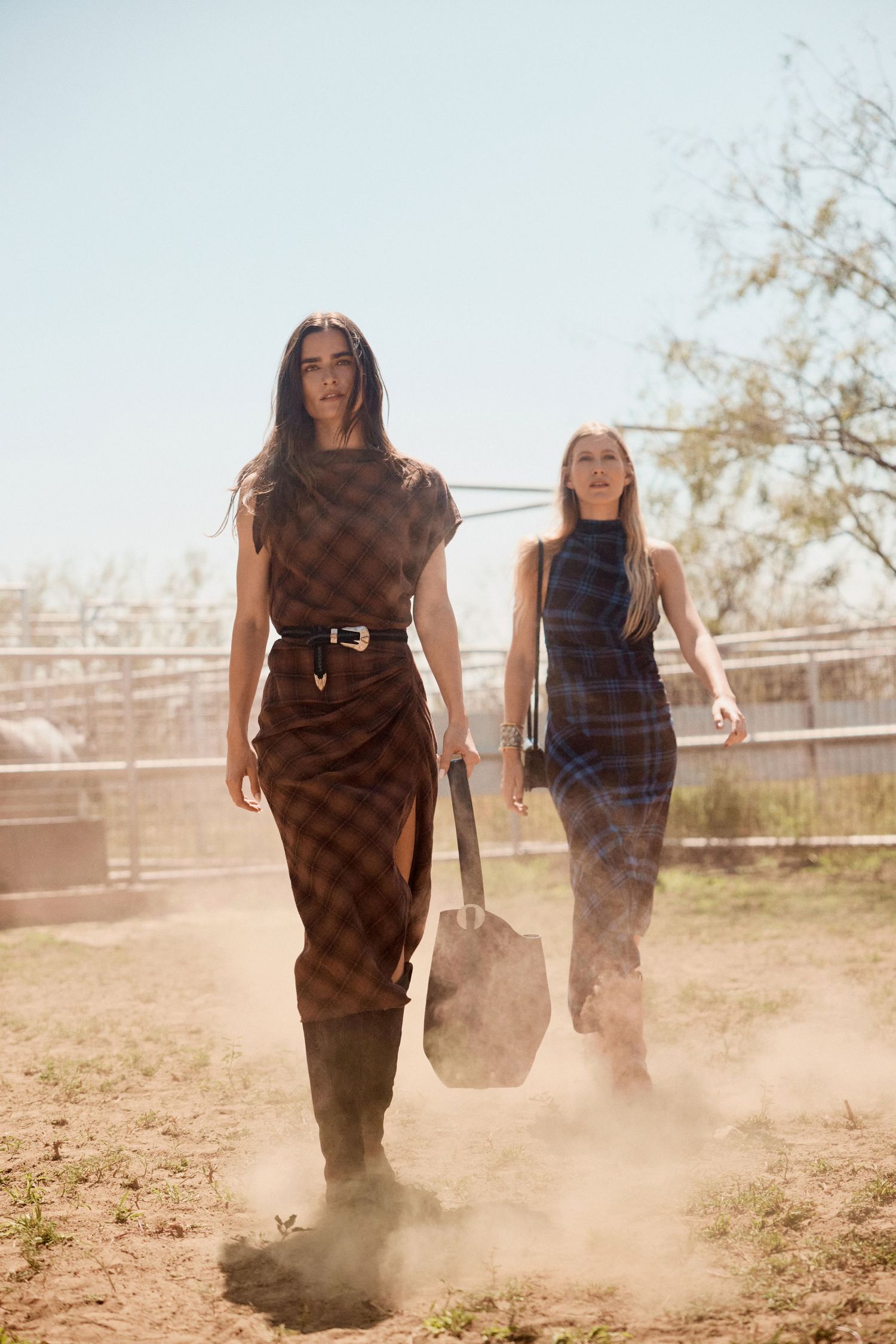 Two women in plaid dresses and boots walk confidently through dusty ranch grounds carrying designer bags.