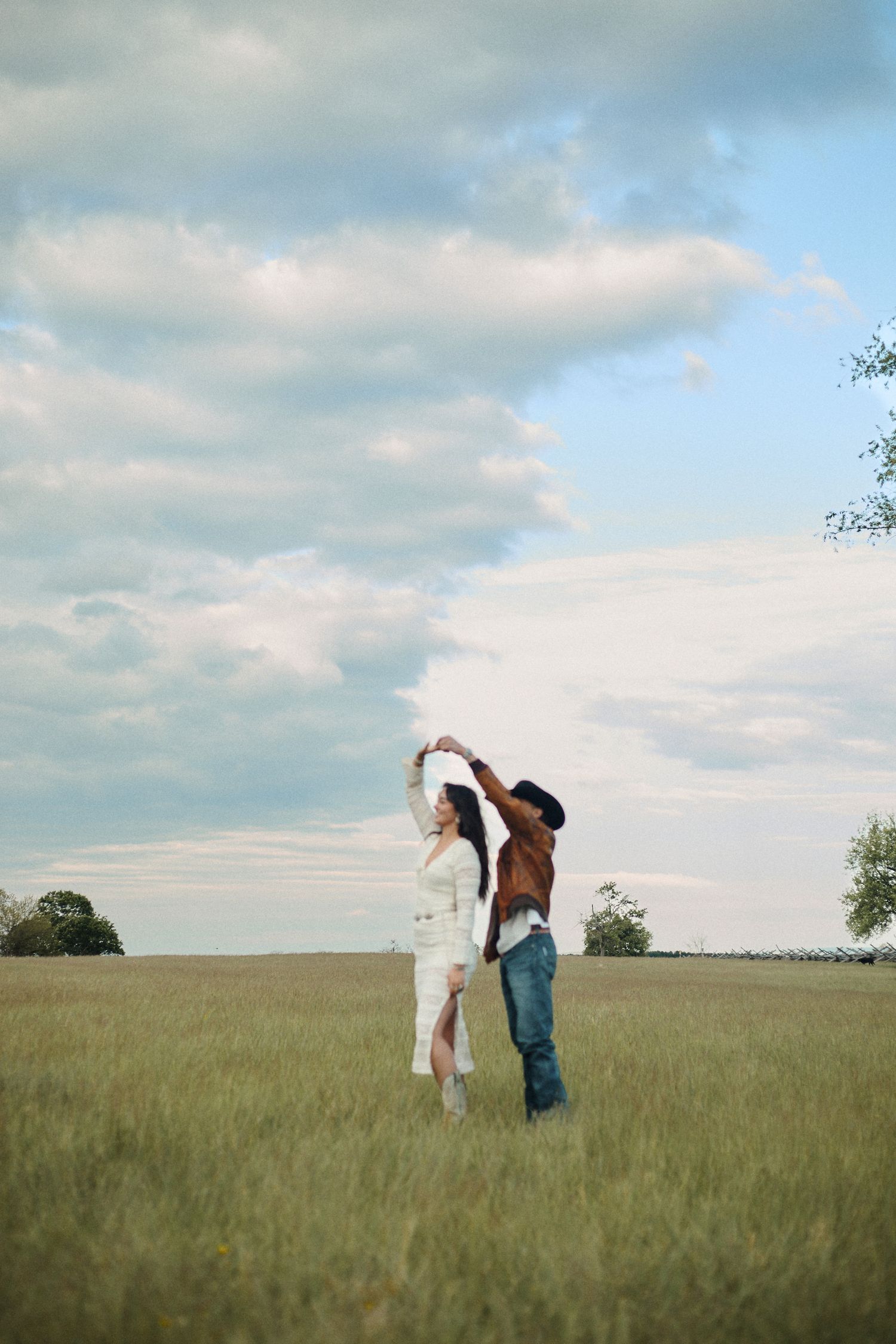 Couple dancing in an open grassy field, woman in white dress, man in cowboy hat and brown leather jacket.