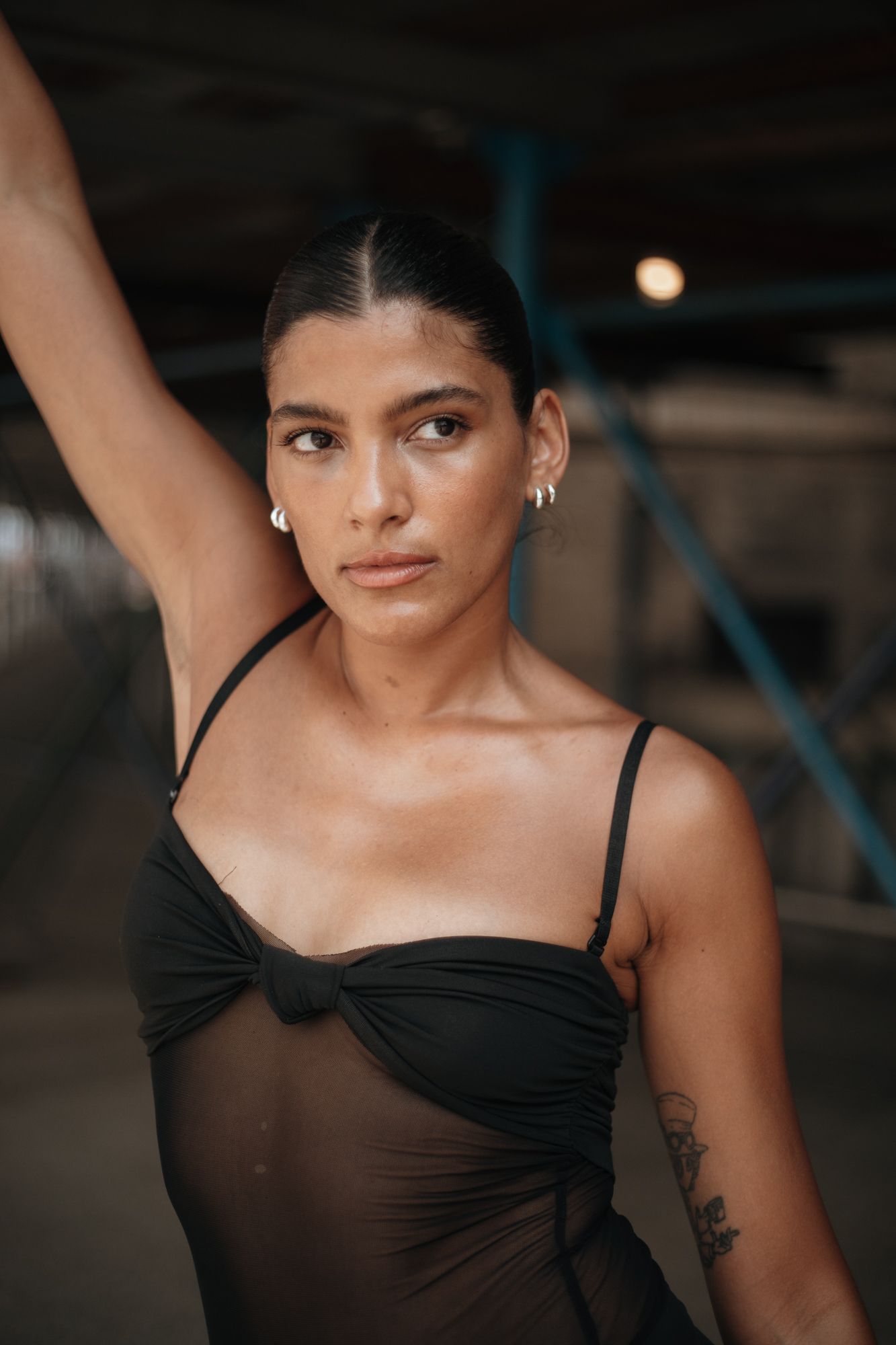 Woman in black sheer dress with tattoo raises arm in a dimly lit urban setting.