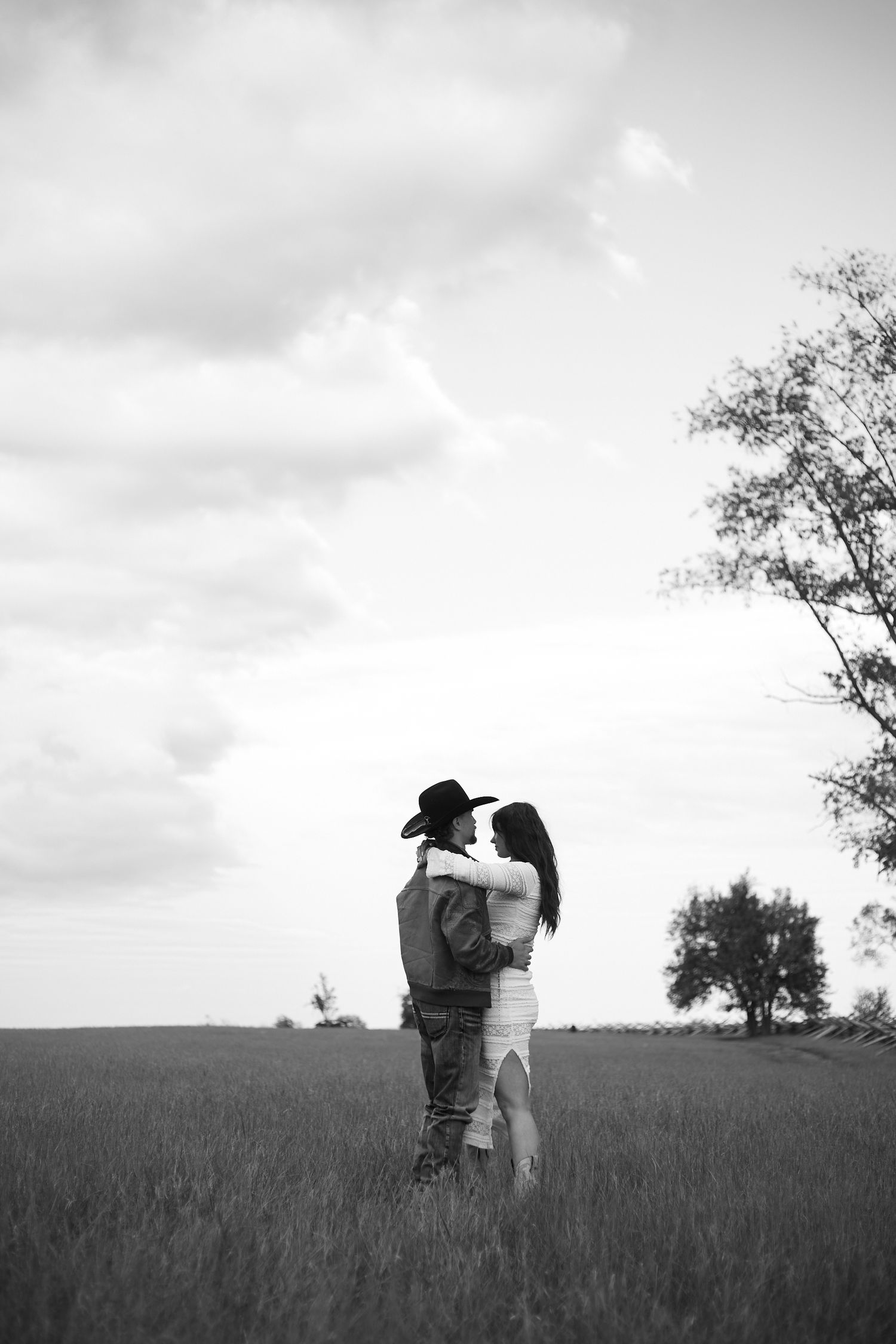 Black and white photo of a couple embracing in a field, man wearing cowboy hat, trees and cloudy sky in background.