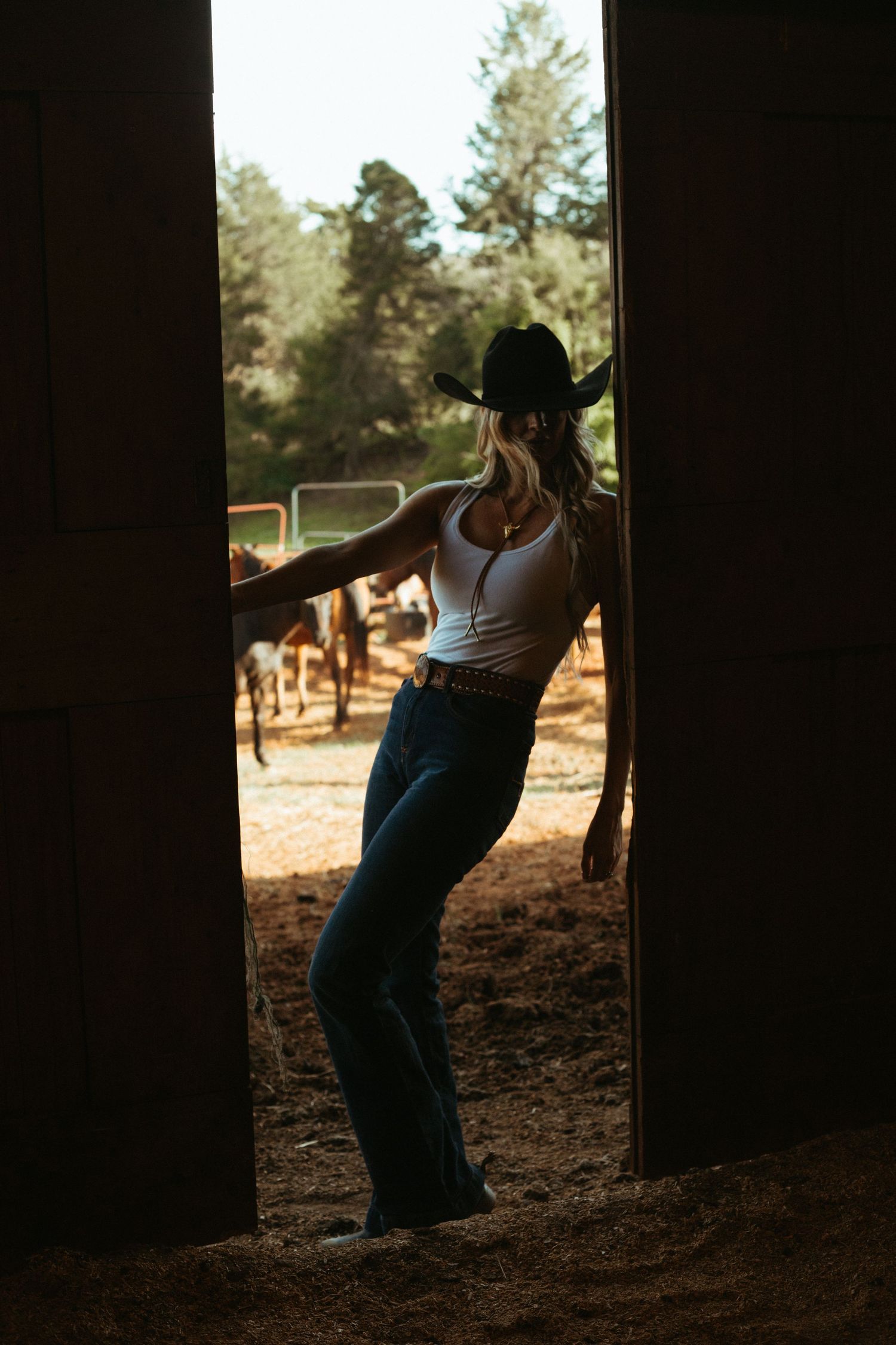 Cowgirl in white tank top, jeans, and black hat stands in barn doorway overlooking horses outside.
