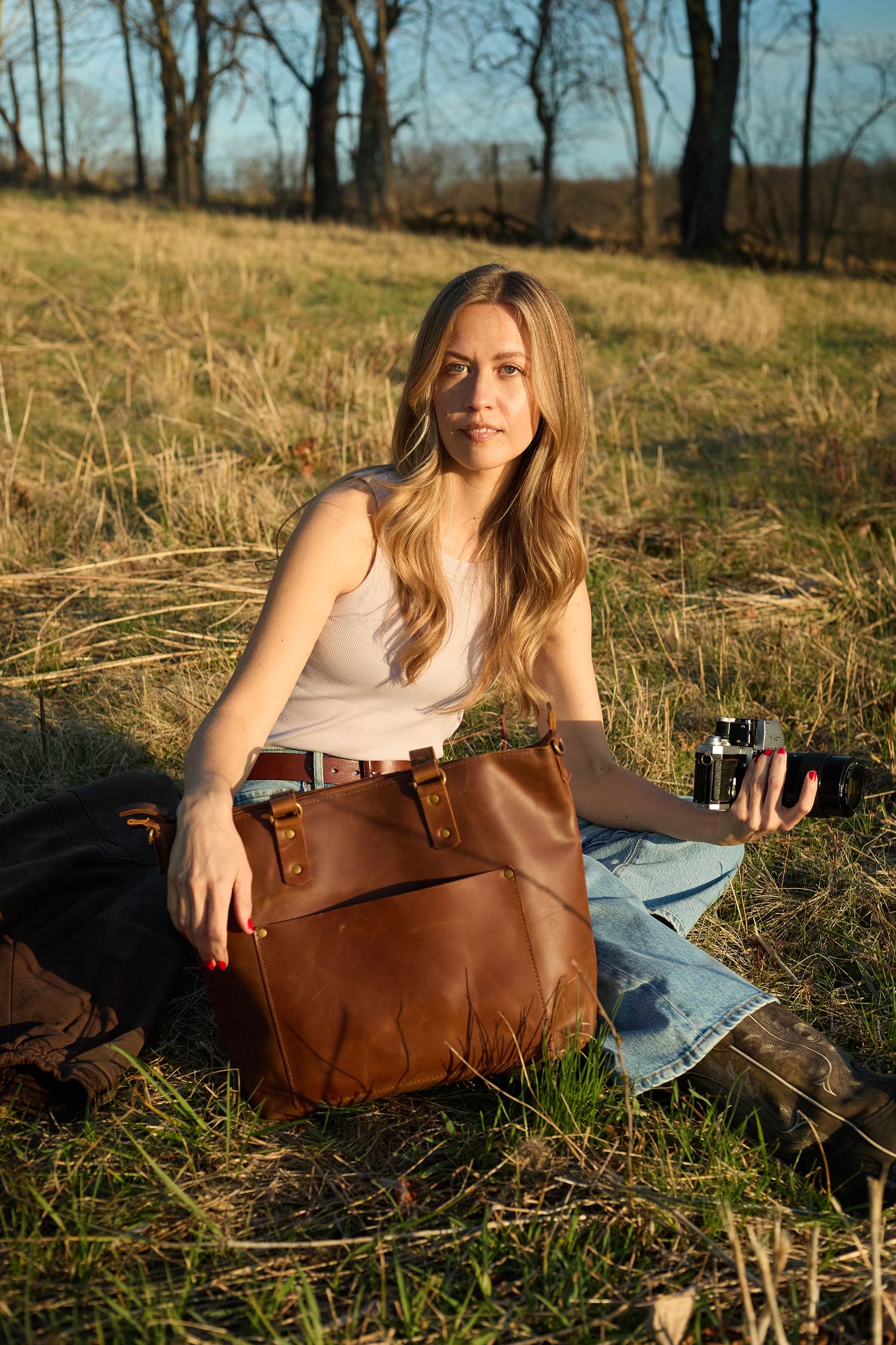 Blonde woman sitting in a dry grassy field with a brown leather bag and vintage camera at sunset.