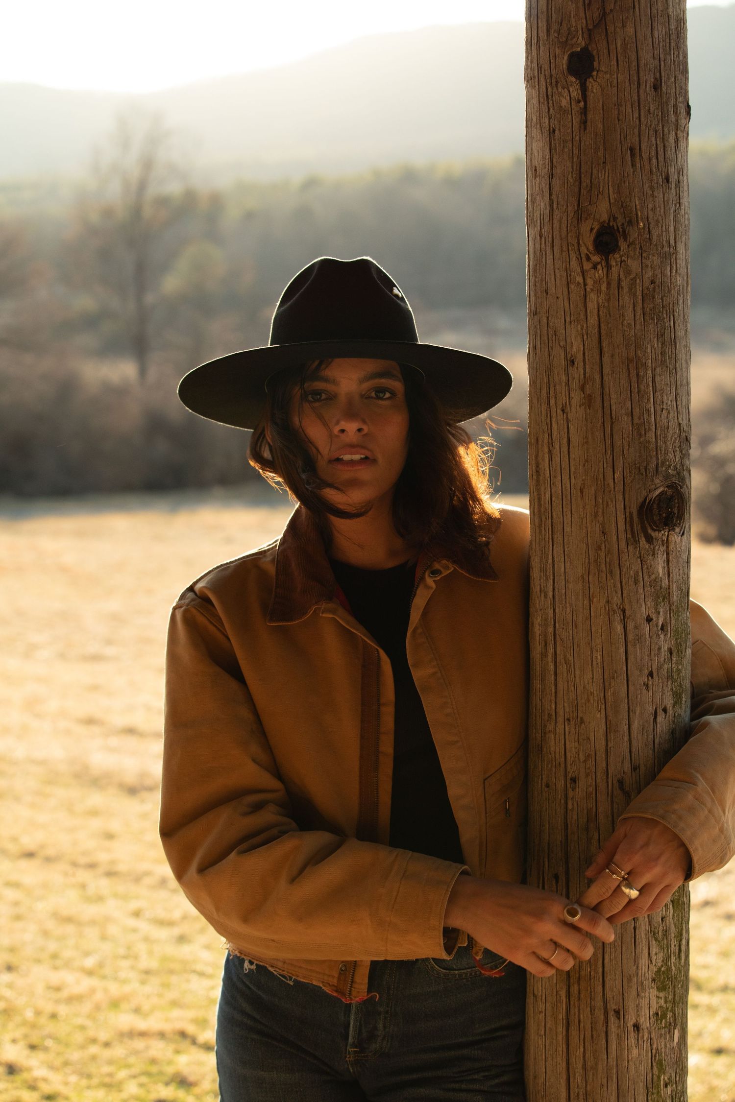 Person in brown jacket and black wide-brim hat leans against a wooden post in a sunlit rural field.