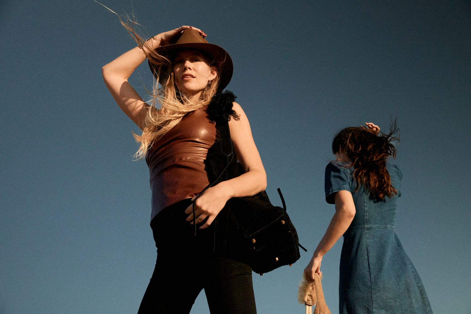 Two women in western-style outfits pose against a clear blue sky, one wearing a cowboy hat.
