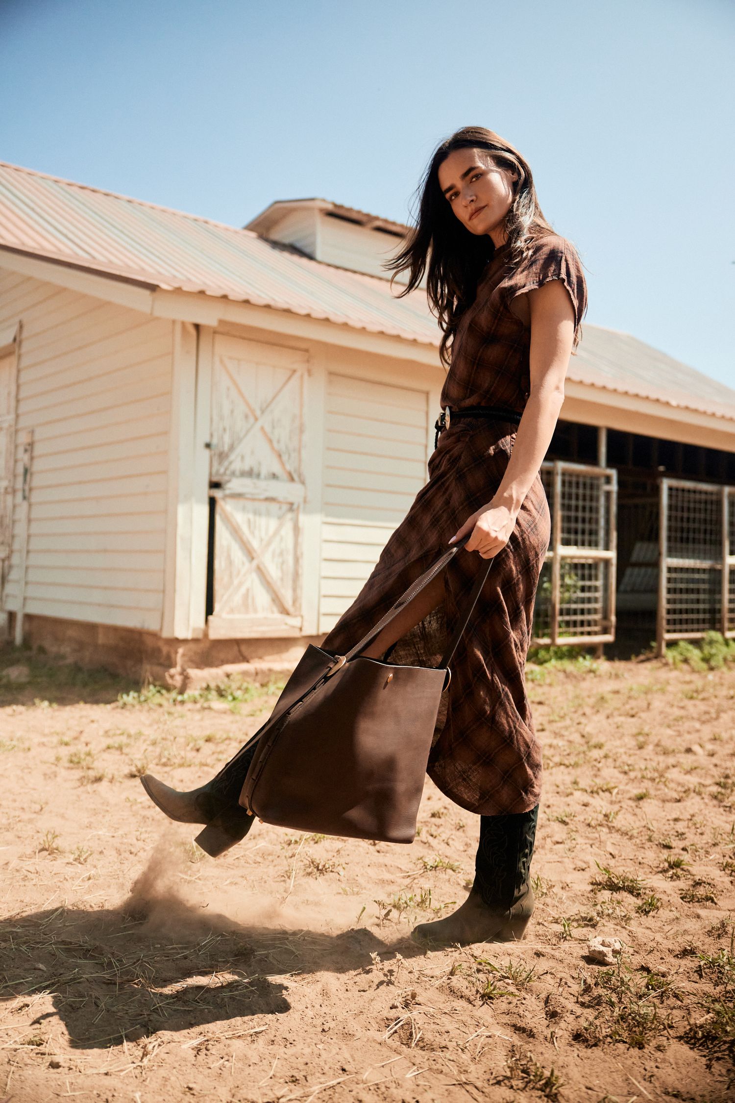 A woman in a brown plaid midi dress carrying a leather tote bag at a rustic farm.