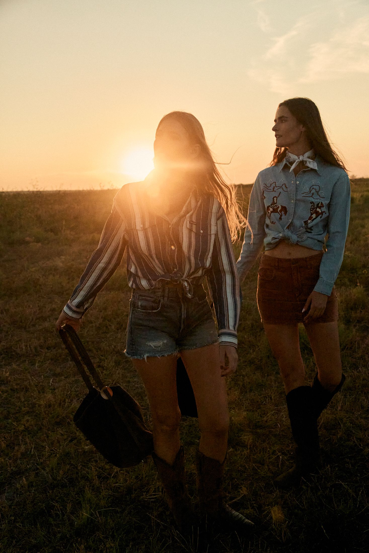 Two women in western outfits with cowboy boots walk through a field carrying bags at sunset.