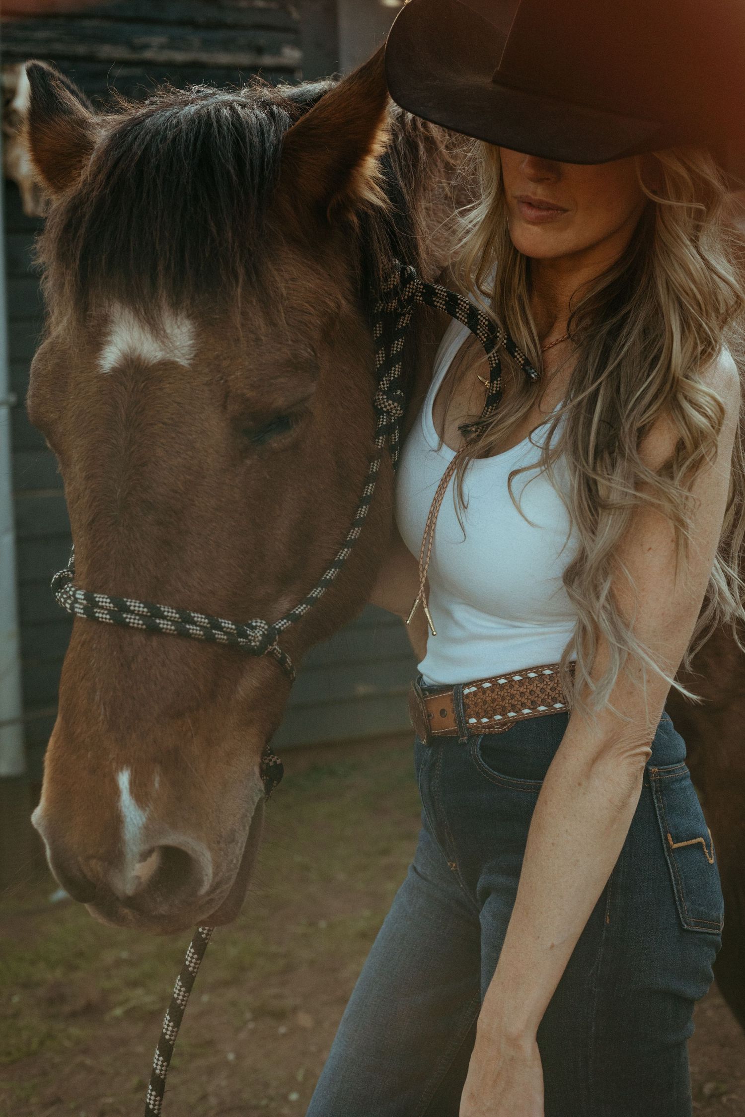 Blonde woman in black cowboy hat and white tank top stands beside a brown horse with a rope halter.